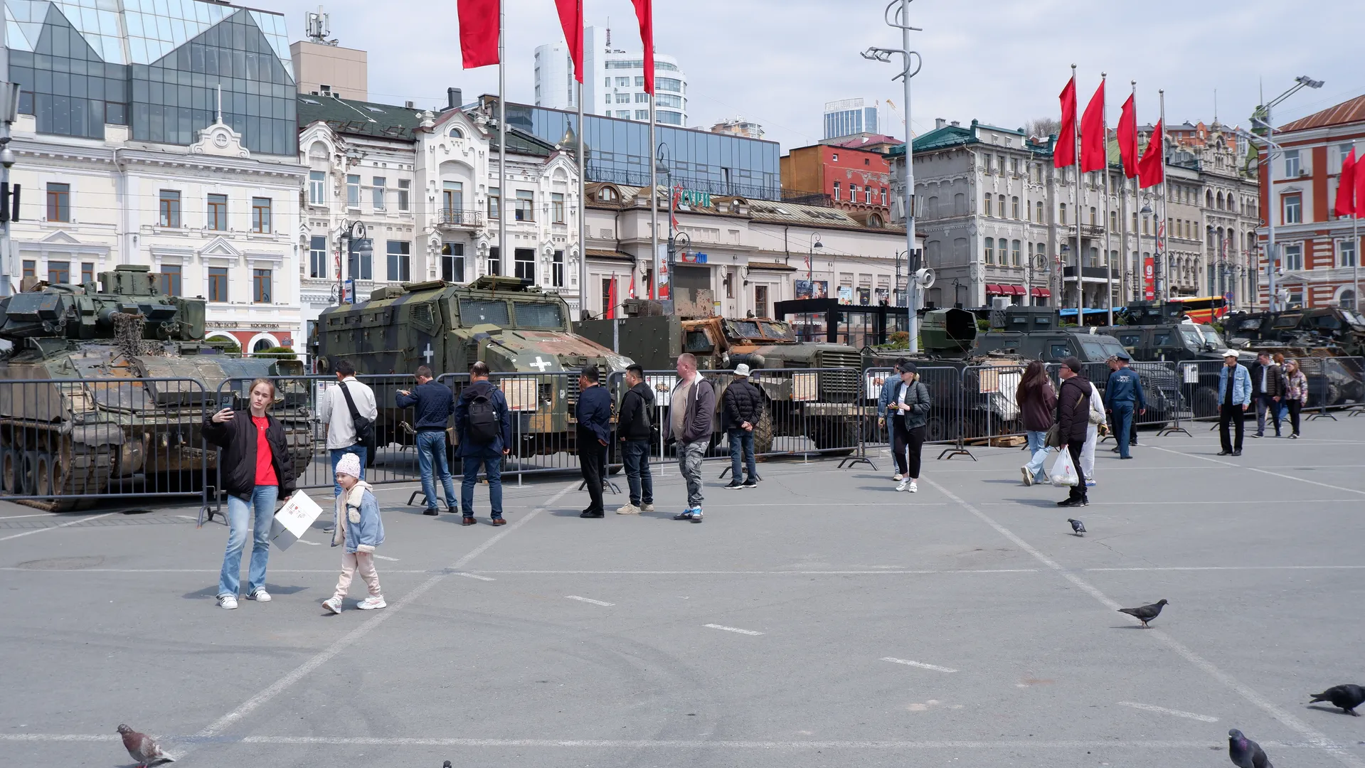 A square displays several military vehicles, surrounded by fences and pedestrians. Background buildings and red flags are visible. The sky is clear and there are several pigeons on the ground.