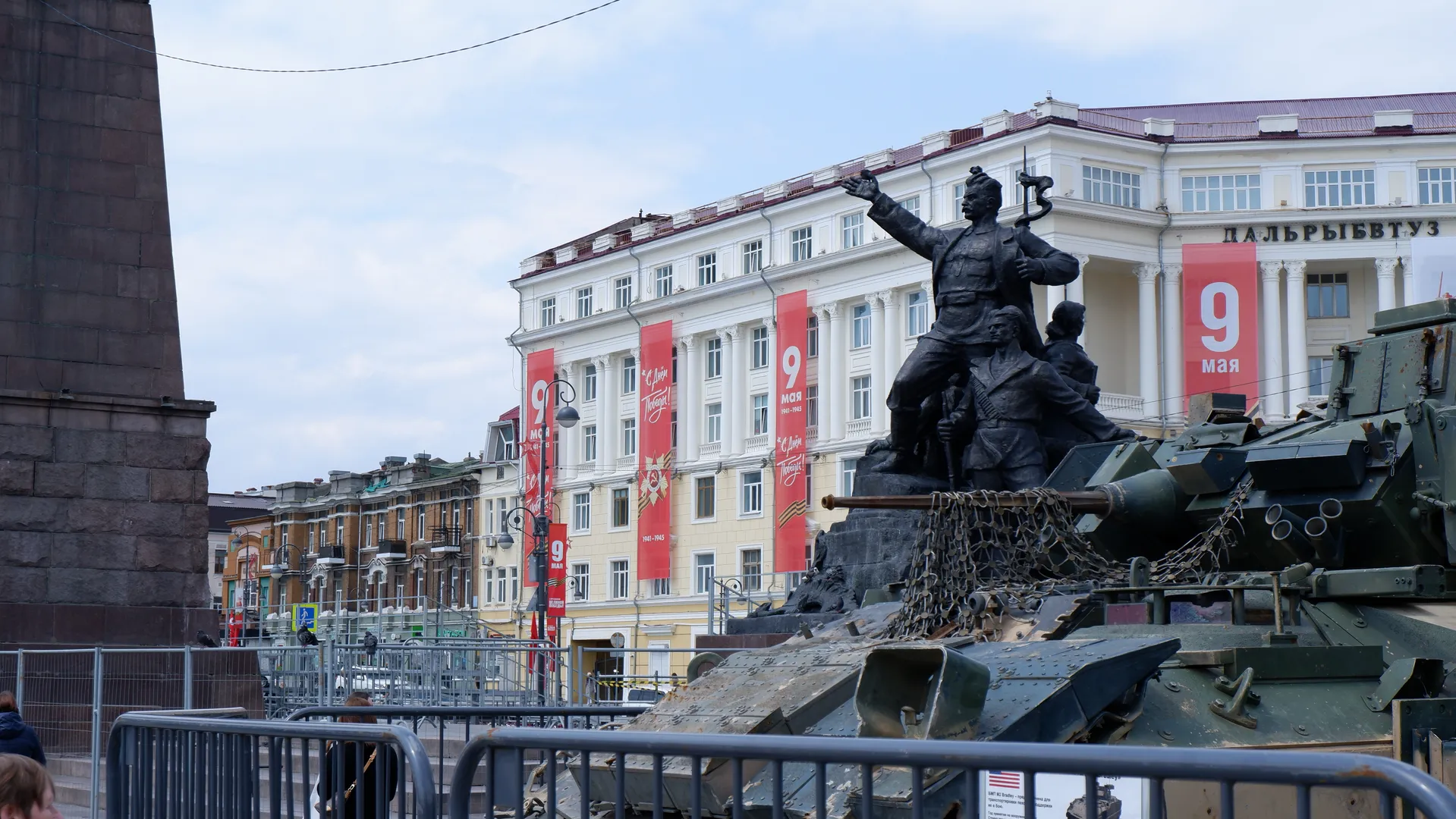 A statue stands on a tank, depicting a heroic figure in motion. The background is a white building with a red banner hanging on it. A military vehicle is parked beside.