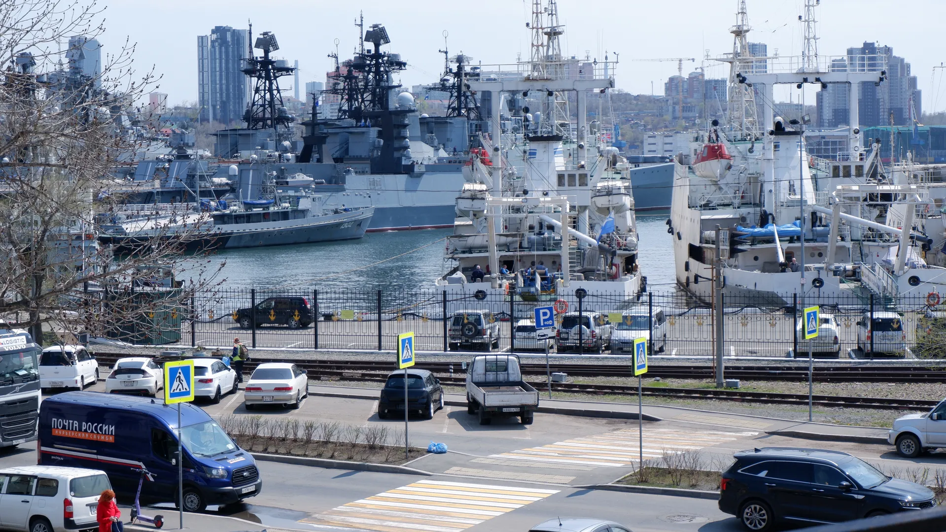 Multiple large ships are docked at the port, with a parking lot and roads in the foreground. Background city buildings and cranes are visible. The sky is clear and the water surface is calm.