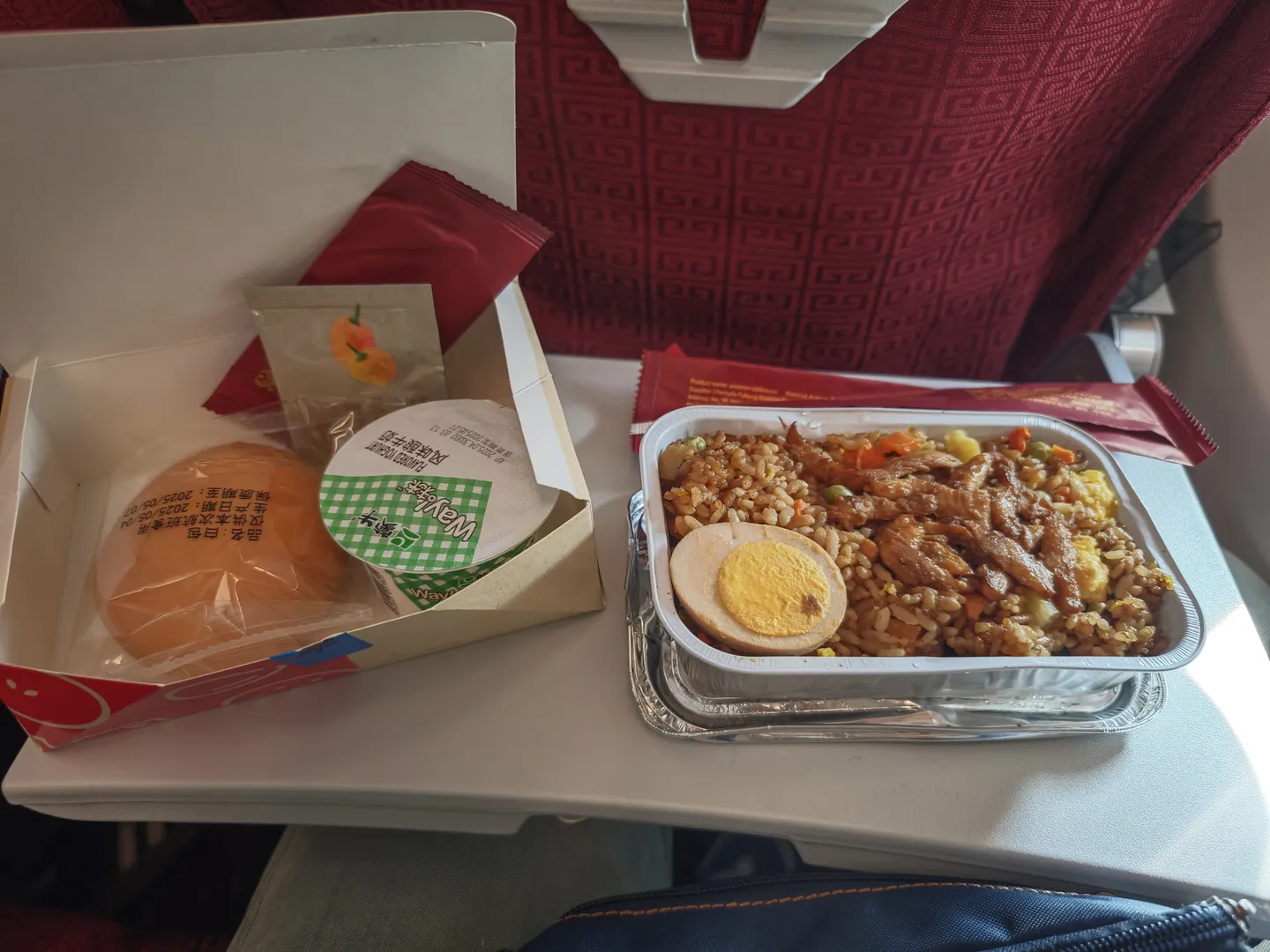 An airplane meal tray sits on a small table, containing fried rice and side dishes. The fried rice includes meat pieces, vegetables, and half a boiled egg. Beside it is a wrapped bread roll.