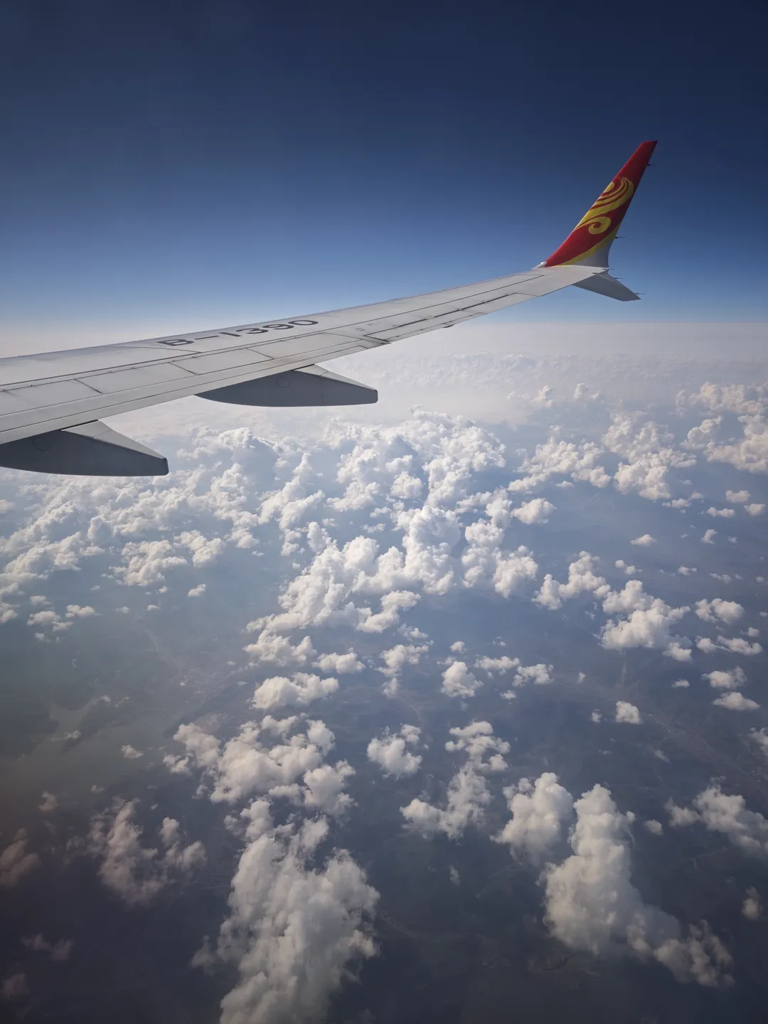 The view outside the airplane window is spectacular, with the wing extending to the right side of the frame. Below are clouds and ground landscapes. The sky is bright blue and the clouds are white.
