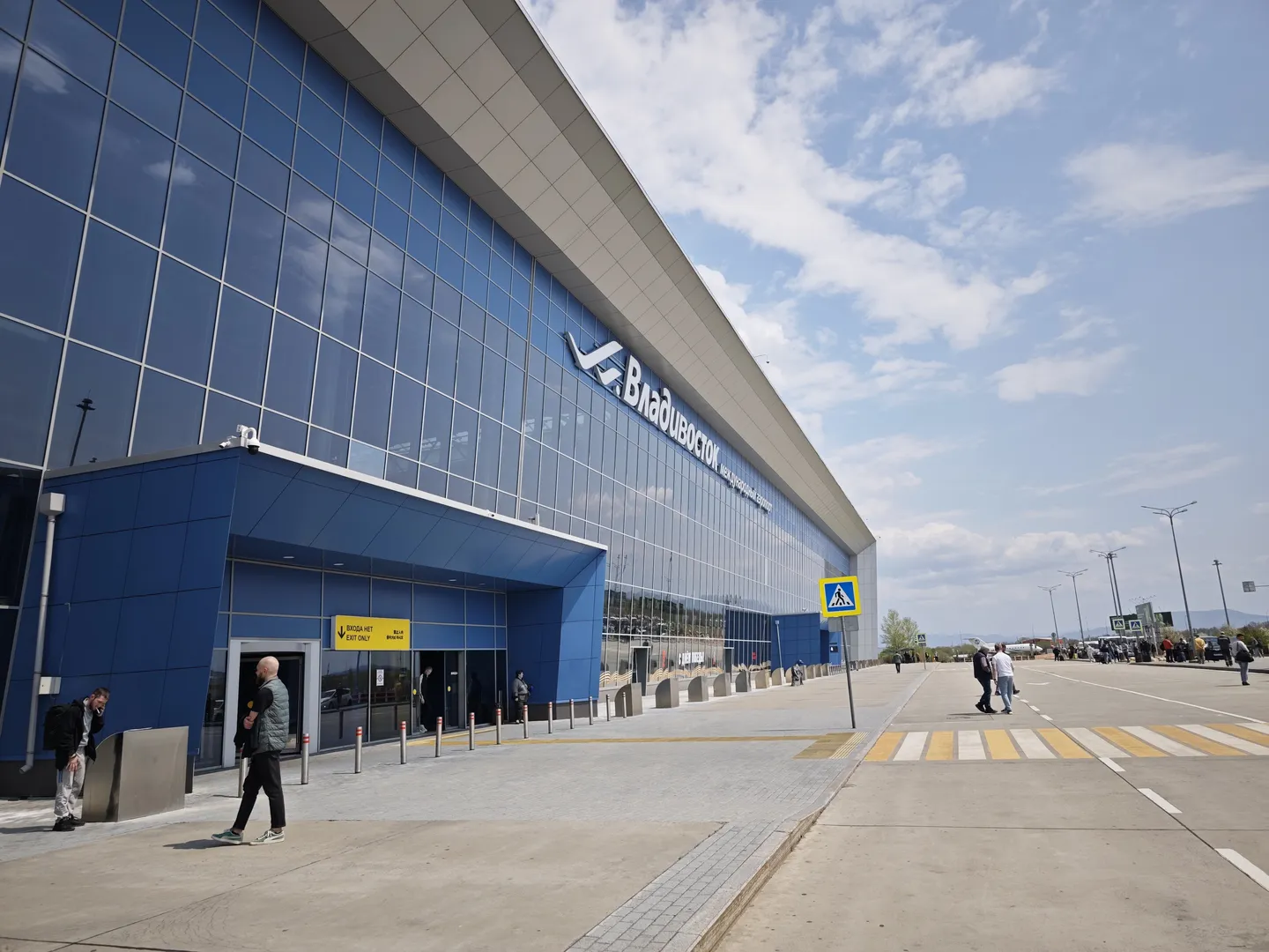 A modern building features a primarily blue glass curtain wall. Several people are walking near the entrance of the building. The sky is clear and the ground area is spacious.