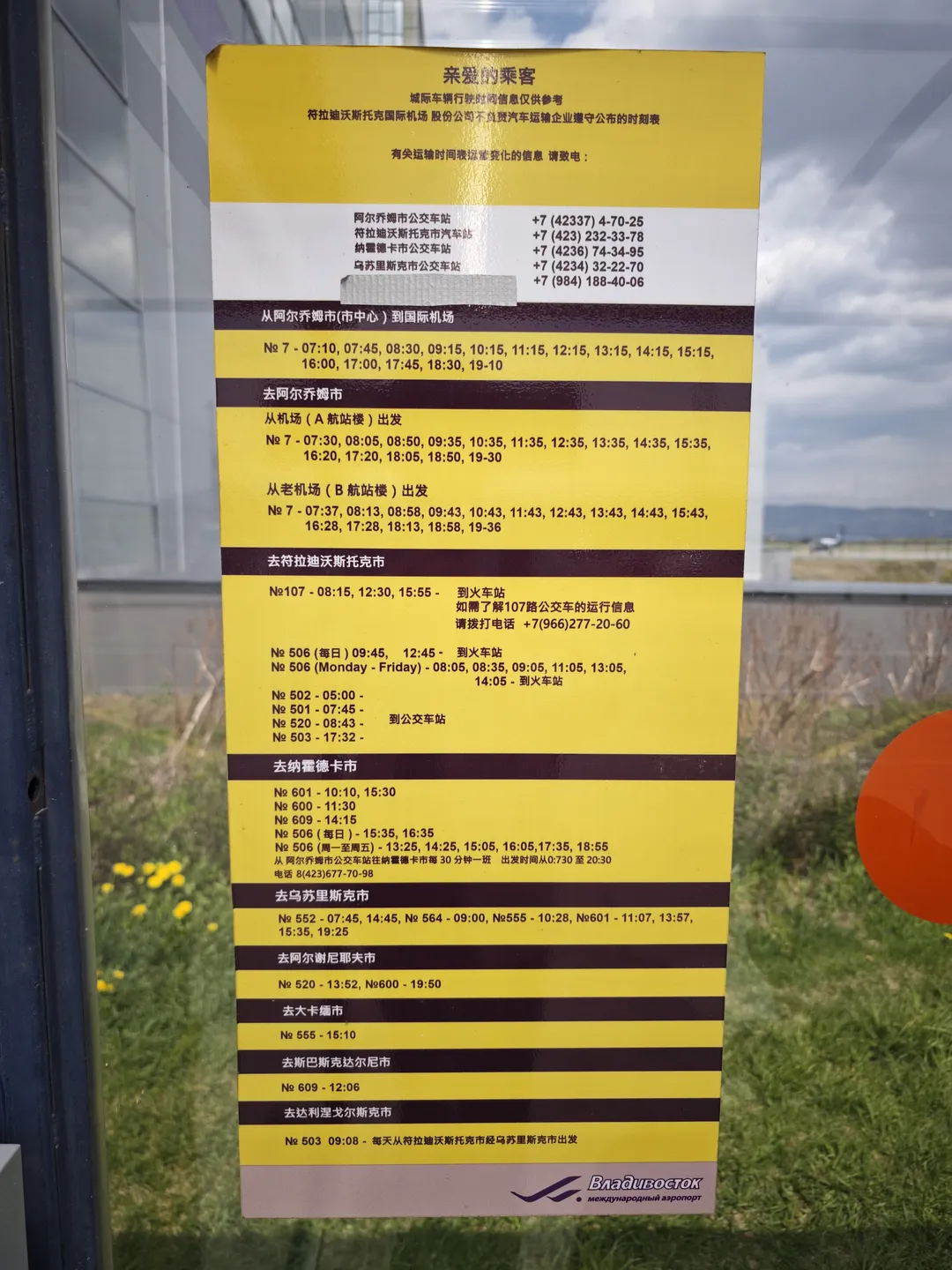 A yellow-background bus timetable is posted on transparent glass. The table details the departure times of different routes. The background shows outdoor grassland and distant mountains.