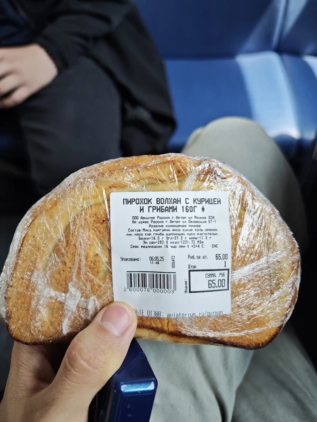A hand holds a round bread roll wrapped in plastic film. Russian text and a barcode appear on the bread’s label. People wearing dark clothes and blue seats are visible in the background.