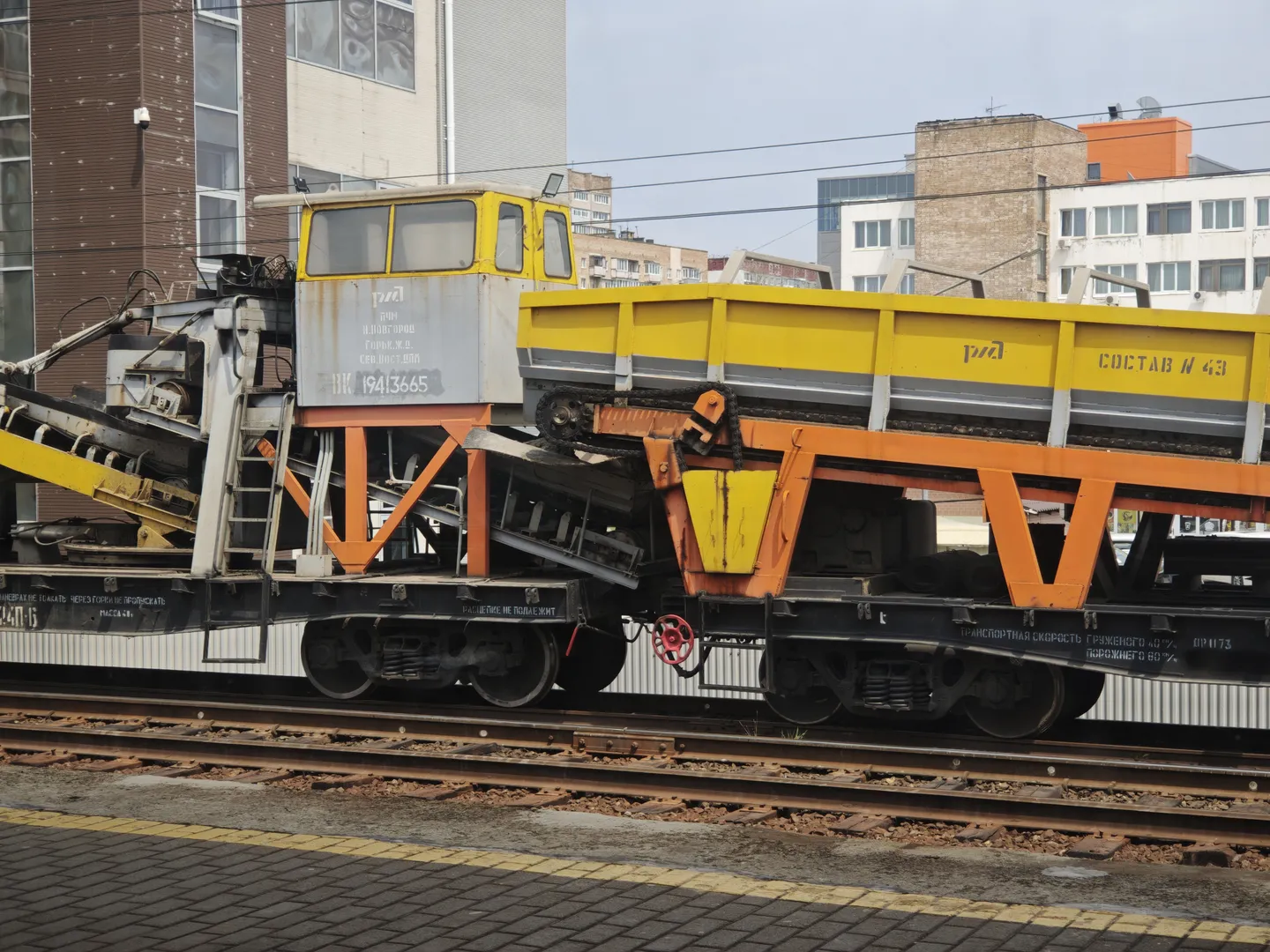 A yellow-and-gray railway maintenance vehicle is parked on the tracks, next to a building. The vehicle has number 19413665 and is connected to an orange trailer. Multi-story buildings are visible in the background.