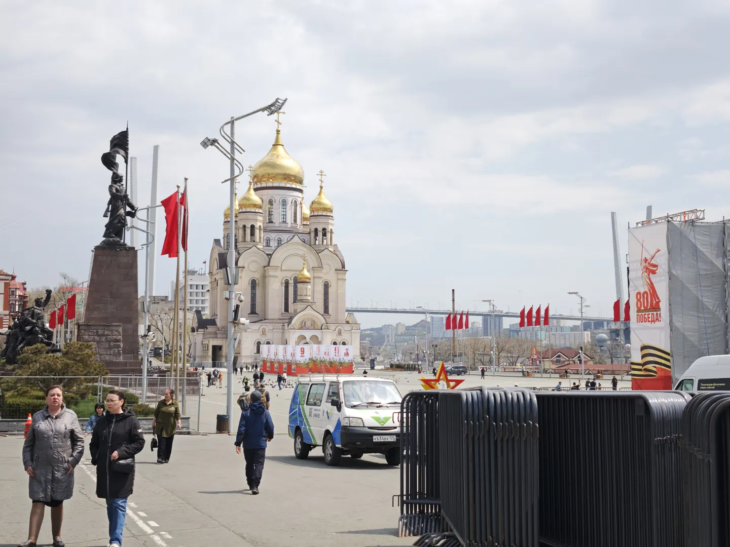 A church with a golden dome stands at the center of the square, with red flags in front. There is a statue on the left side of the church and a propaganda signboard on the right. Pedestrians and vehicles are present in the square.
