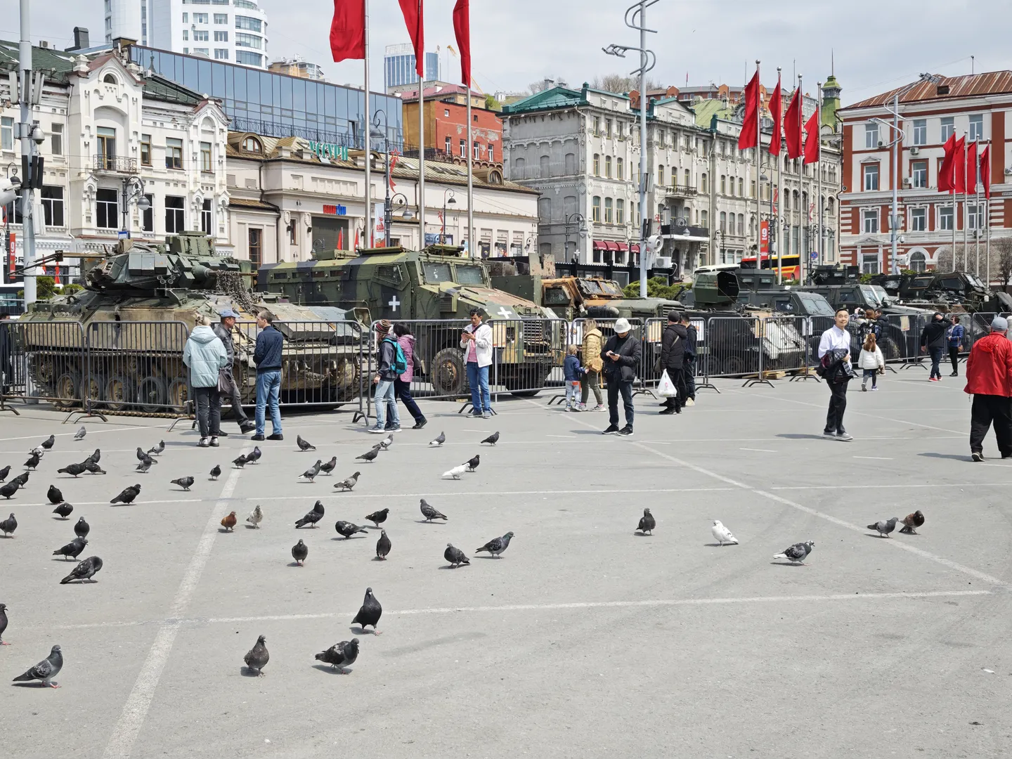 Several military armored vehicles are parked in the square, surrounded by fences. There are many pigeons on the ground, and people are walking near the armored vehicles. The background consists of multi-story buildings and red flags.