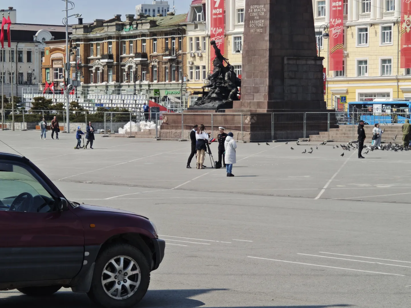 A large sculpture is located at the center of the square, surrounded by fences. Several people are taking photos in front of the sculpture, and pigeons are on the ground. The background consists of multi-story buildings and red flags.