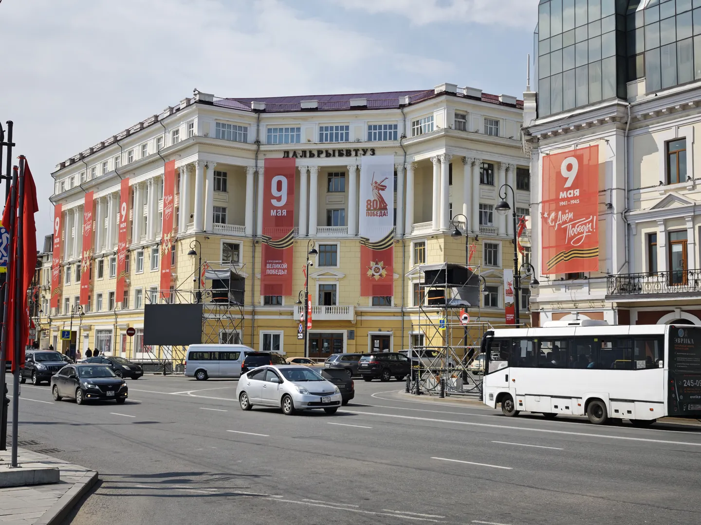 Buildings along the street are decorated with red banners bearing the words “9 мая”. Cars and buses are driving on the road. The architectural style is classical with neatly arranged windows.