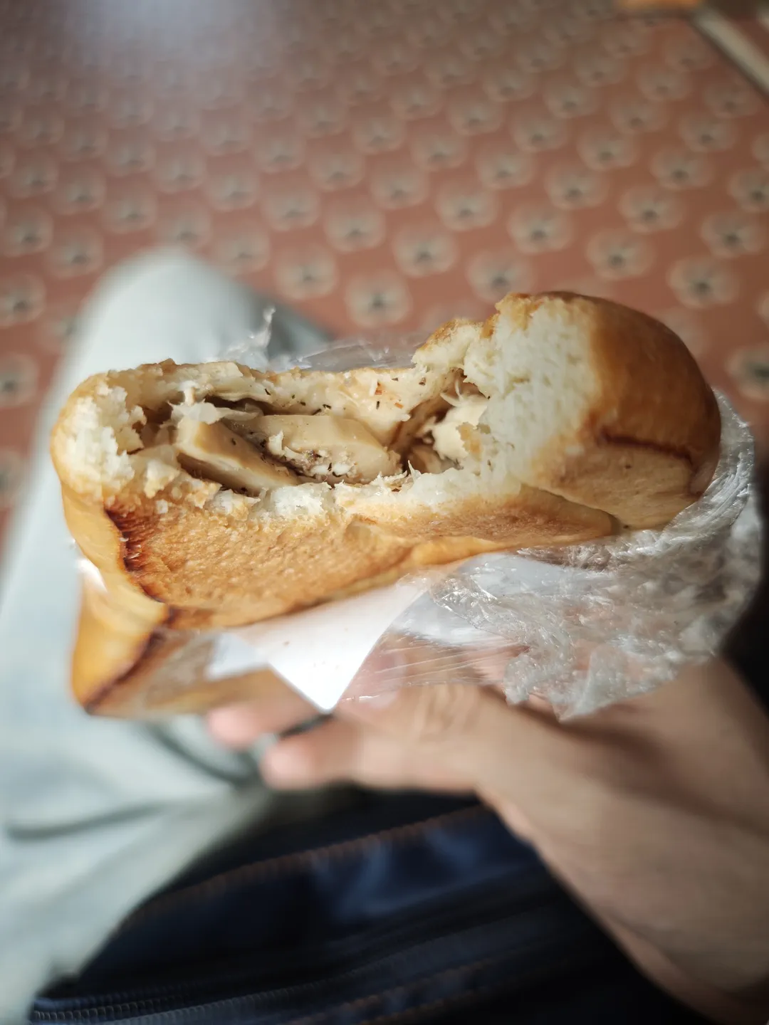 A hand holds a partially eaten bread roll filled with cream-like content. The bread crust is golden and wrapped in a transparent plastic bag. The background features a floor with circular patterns.