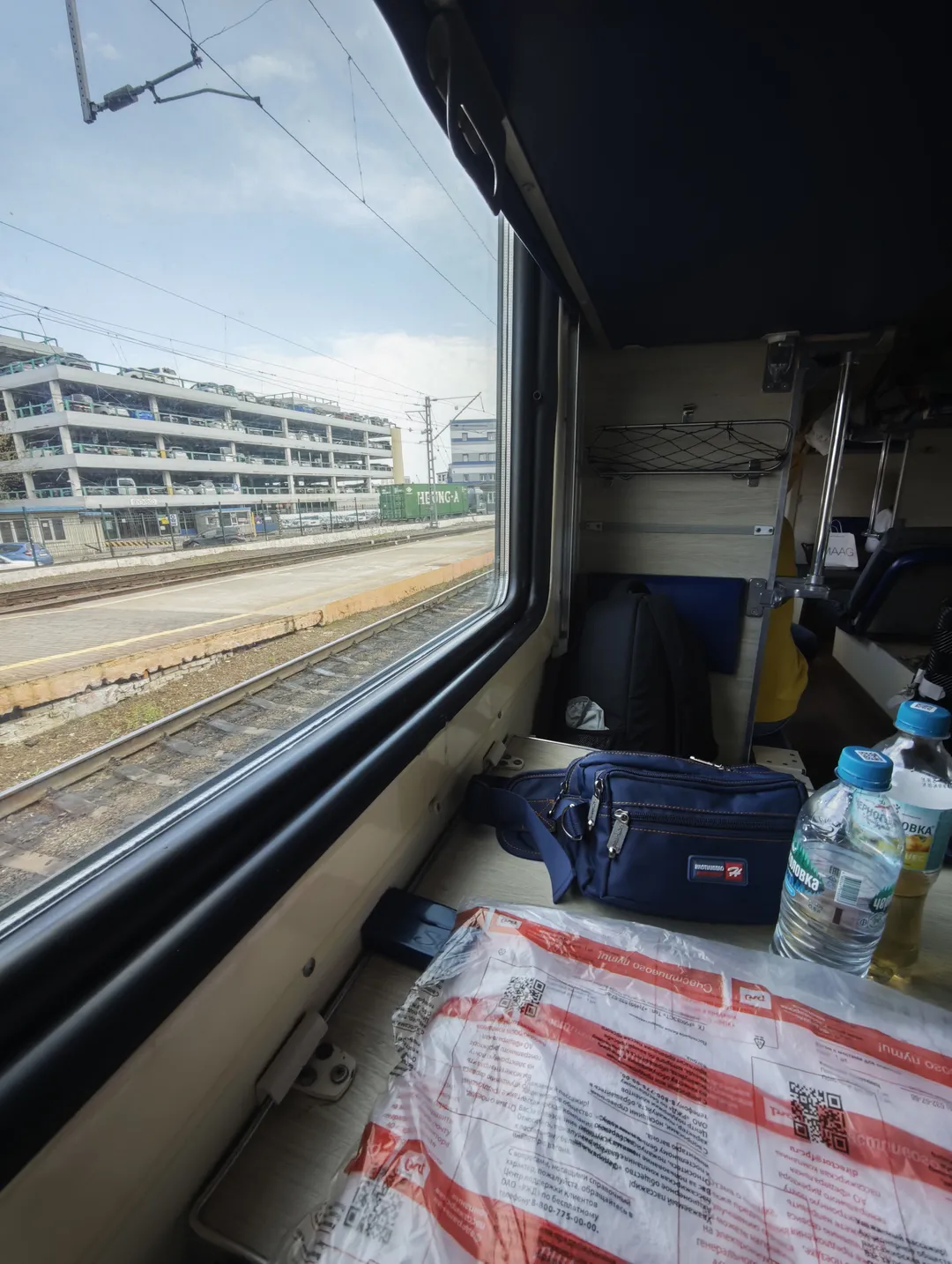 Inside a train carriage, the view outside reveals railway tracks and a multi-level parking lot. A blue bag and water bottle are placed near the window. Luggage racks and seats are present inside the carriage.