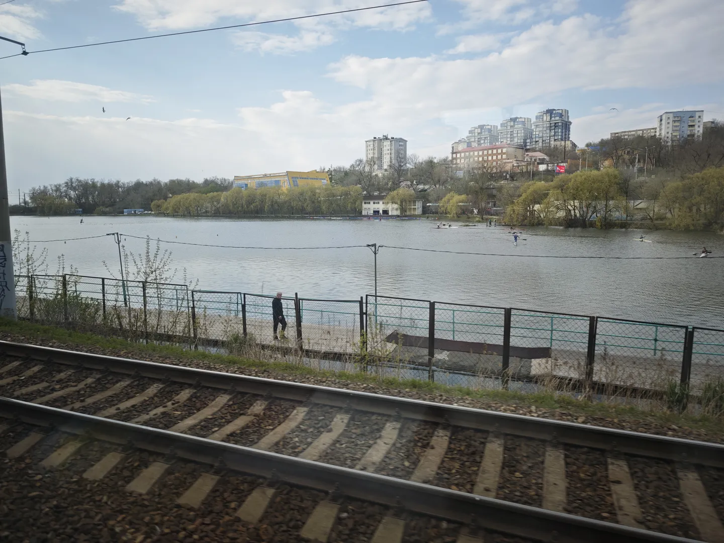 Beside the railway tracks lies a river with people rowing boats on it. Trees and buildings line the riverbanks, with high-rise buildings visible in the distance. Clouds are present in the sky, and power lines cross the scene.