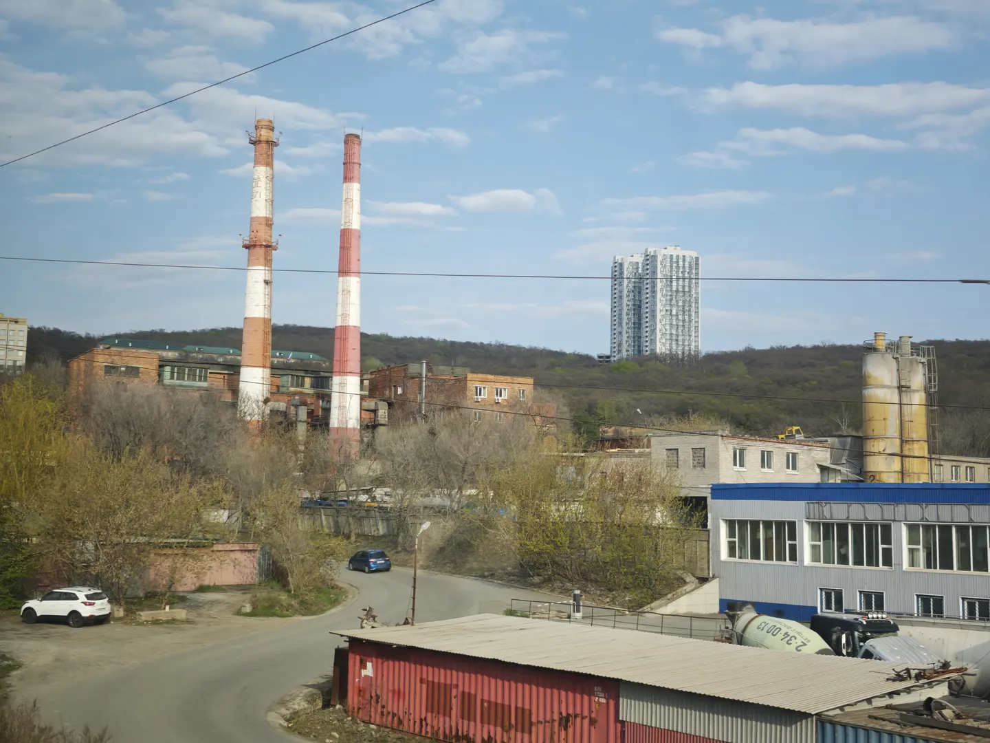 An industrial area scene featuring two tall chimneys in red and white stripes. The foreground includes a road and parked vehicles, while the background consists of trees and high-rise buildings. Industrial facilities coexist with residential buildings.
