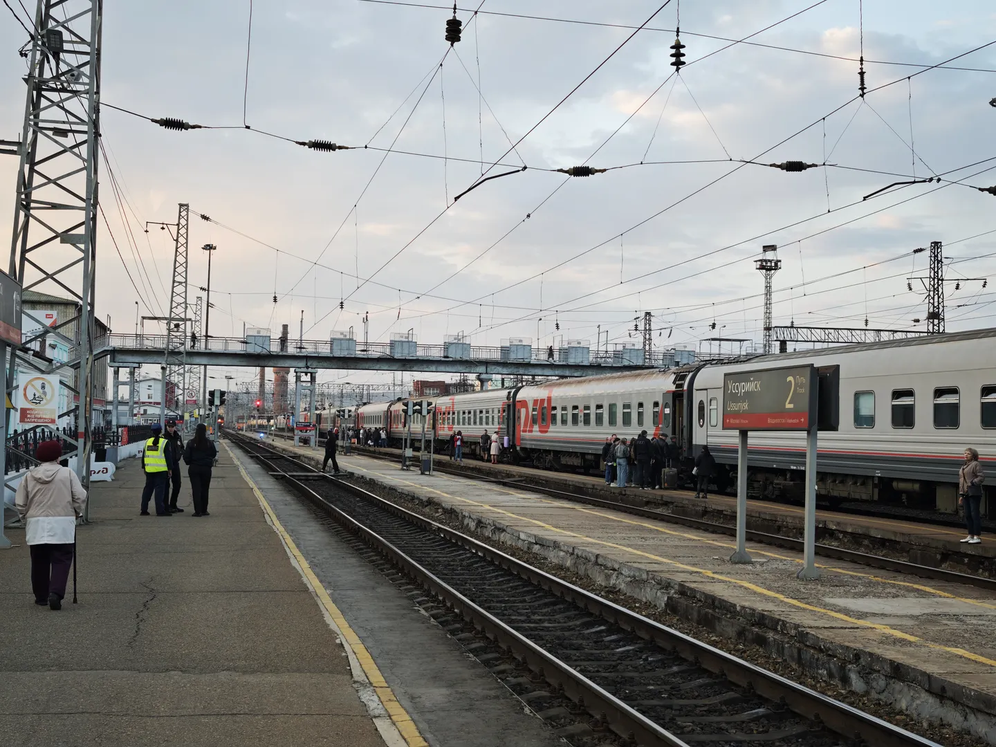 A train platform with multiple railway tracks and a stopped train. Crowds gather on the platform, with overhead wires and signal lights above. A footbridge connects the platforms in the distance.