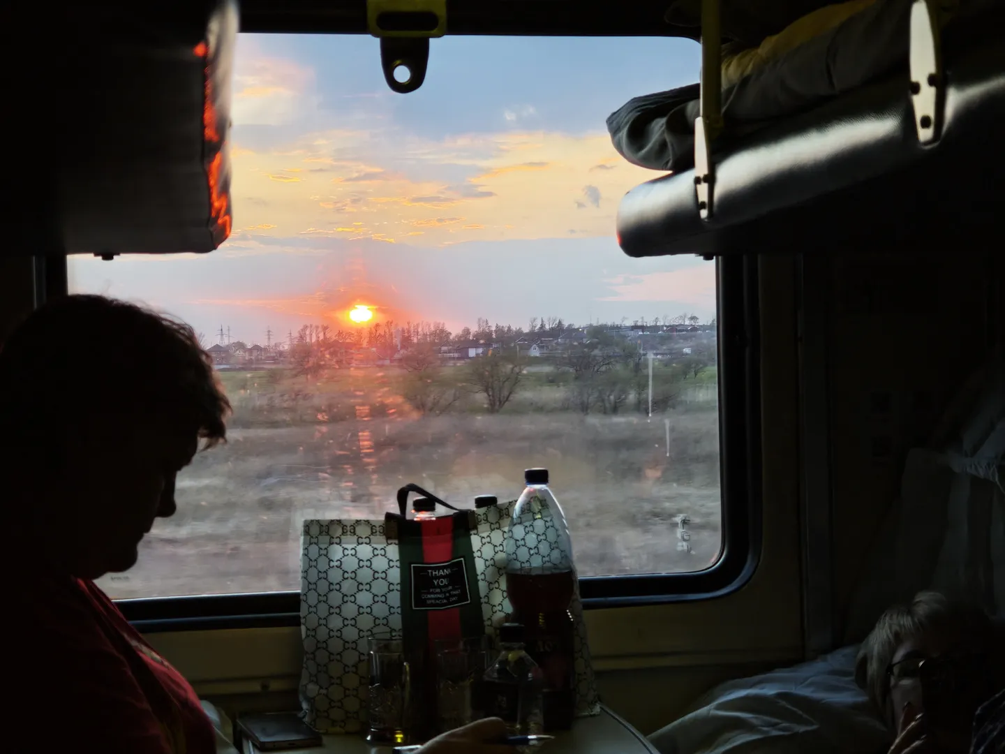 The sunset can be seen through the window as it slowly descends, painting the sky in shades of orange and red. Inside the cabin, a person sits sideways, with a beverage and a shopping bag displaying a logo placed in front. On the opposite bunk, someone is lying down beside some belongings.