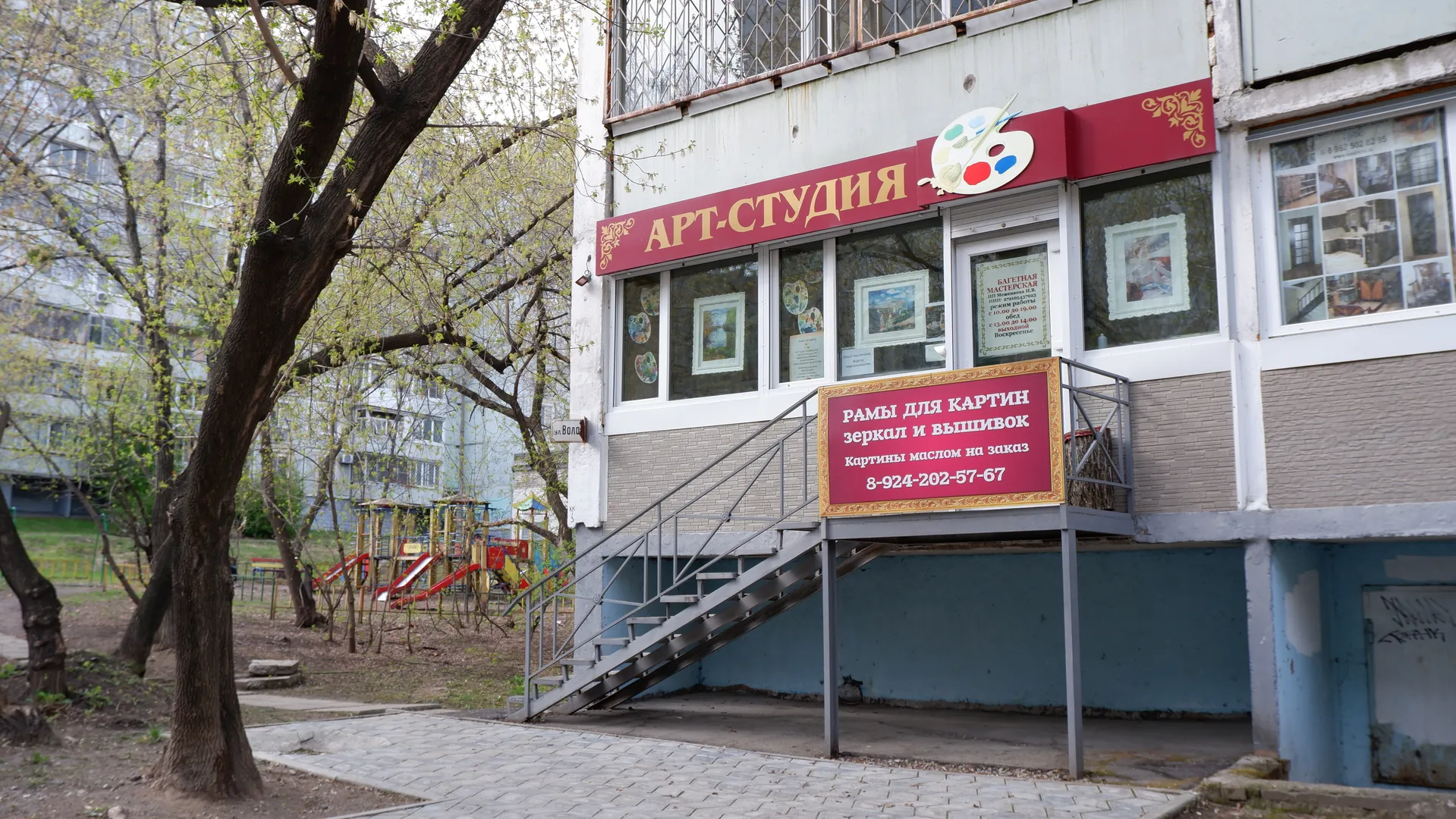 The exterior of an art studio, with a red sign reading “АРТ-СТУДИЯ”. There are stairs and displayed paintings at the entrance. Next to it are trees and a children’s playground with colorful equipment. The building’s facade is pink and white, with shop signs on the ground floor and several cars parked in front.