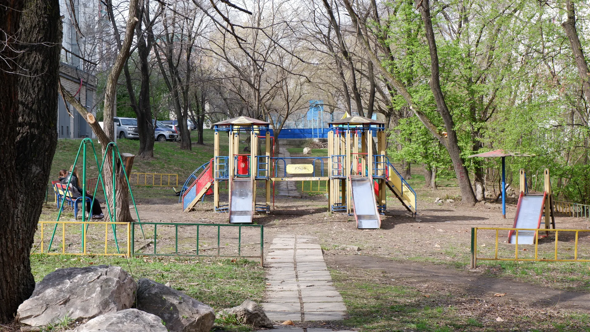 Inside a children’s playground, there are slides and climbing frames, surrounded by trees and yellow fencing on the ground. An adult sits near a swing, and several cars are parked in the distance. The ground is paved with bricks, with steps and a building nearby.