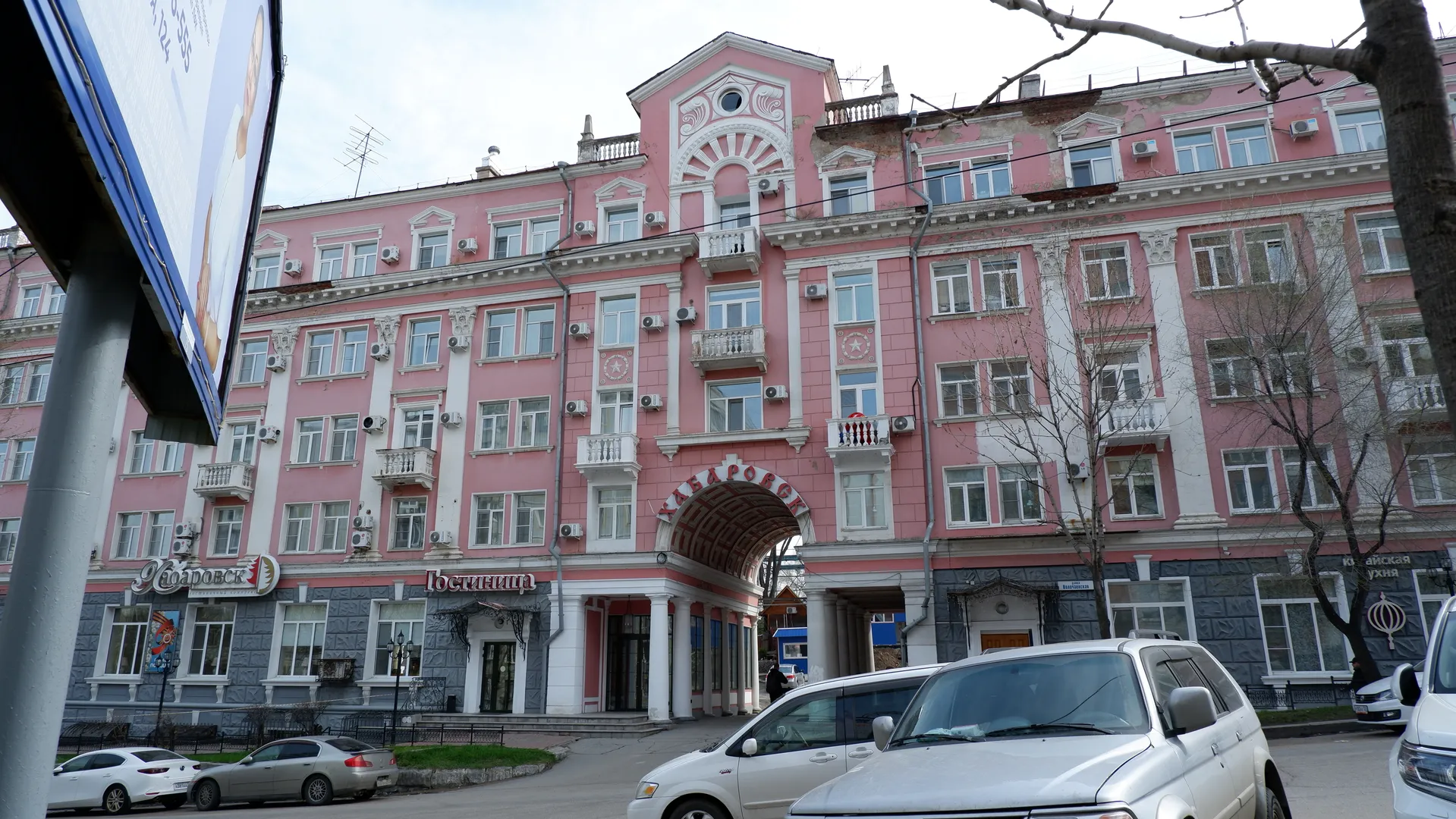 A multi-story building with a light-colored brick exterior, arched porches and shop signs on the ground floor. Several cars are parked in front of the building under a clear sky. The building features a classical architectural style with neatly aligned windows.