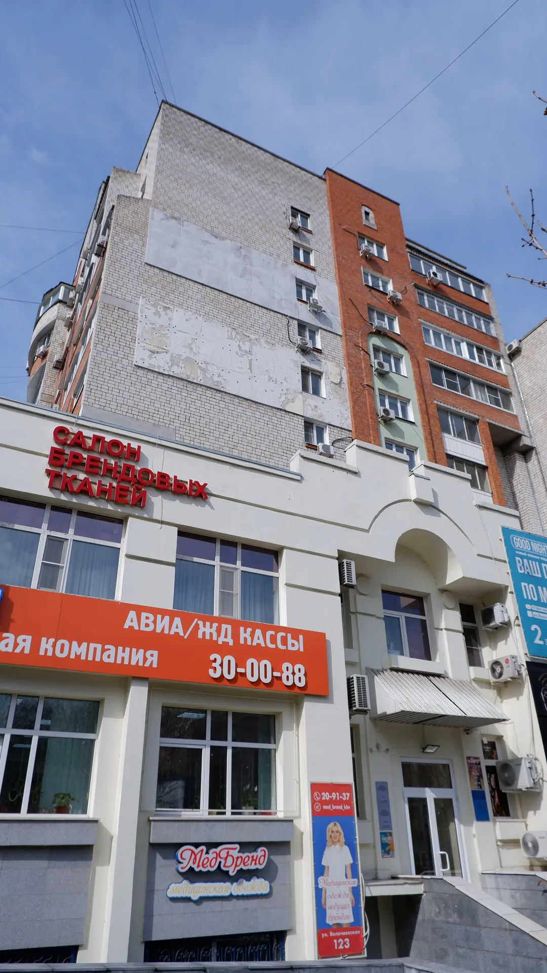 A high-rise building with a ground-level shop sign that reads “САЛОН БРЕНДОВЫХ ТКАНЕЙ”. Parts of the exterior wall are peeling, and the windows are neatly aligned. The sky is clear, with power lines crossing the scene.