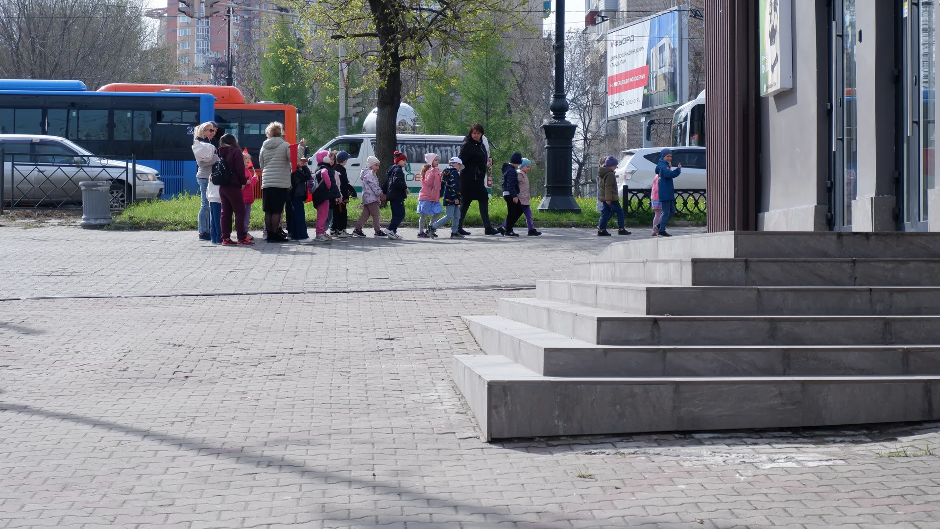 A group of people walking on a plaza with buses and trees in the background. The plaza is paved with stone bricks, with steps and buildings nearby. People are dressed in spring clothing, and the scene is busy.