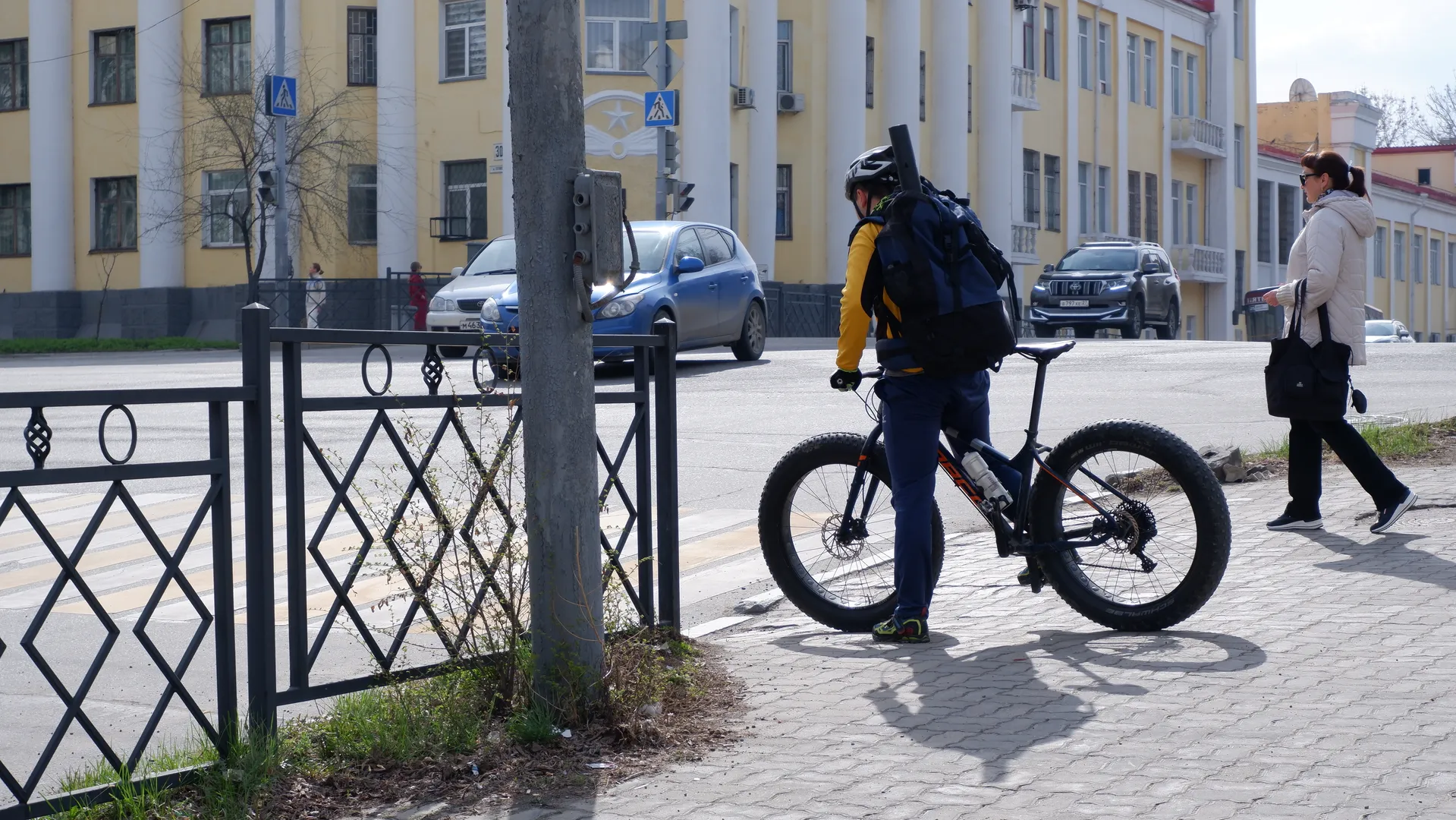 A person with a bicycle stands by the roadside next to a pedestrian. In the background is a yellow building with white columns. Vehicles are driving on the street, and traffic lights are clearly visible.