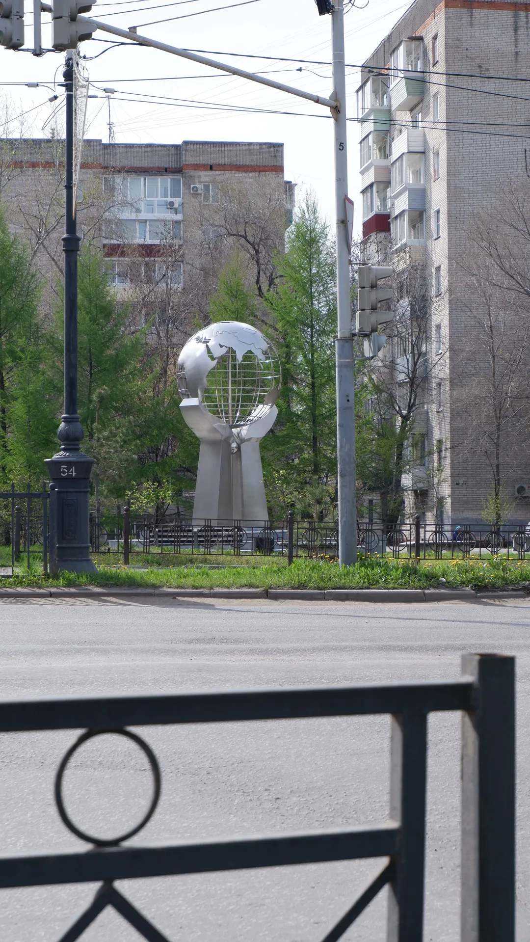 A silver globe sculpture is located in the middle of the road, surrounded by greenery and trees. Behind the sculpture is a high-rise building with neatly aligned windows. There are traffic lights and street lamps on the road.