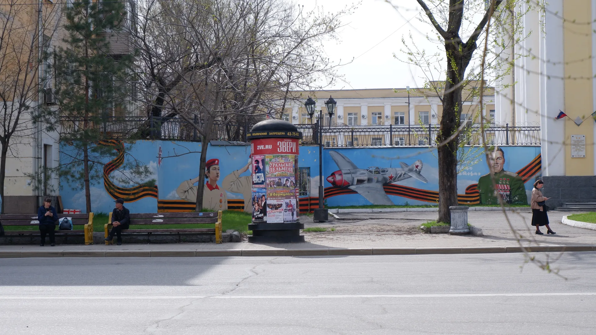 A blue mural featuring soldiers and airplanes, with benches and pedestrians in front. Next to the mural is a yellow building with white columns. The scene also includes trees and billboards.