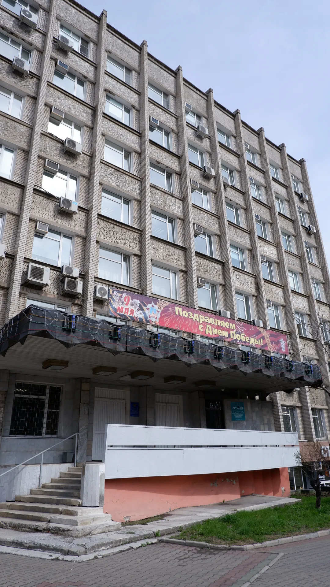 A multi-story building with entrance steps and red decorations on the ground floor. A banner celebrating Victory Day hangs on the front, and the windows are neatly arranged. The sky is overcast with greenery along the ground.