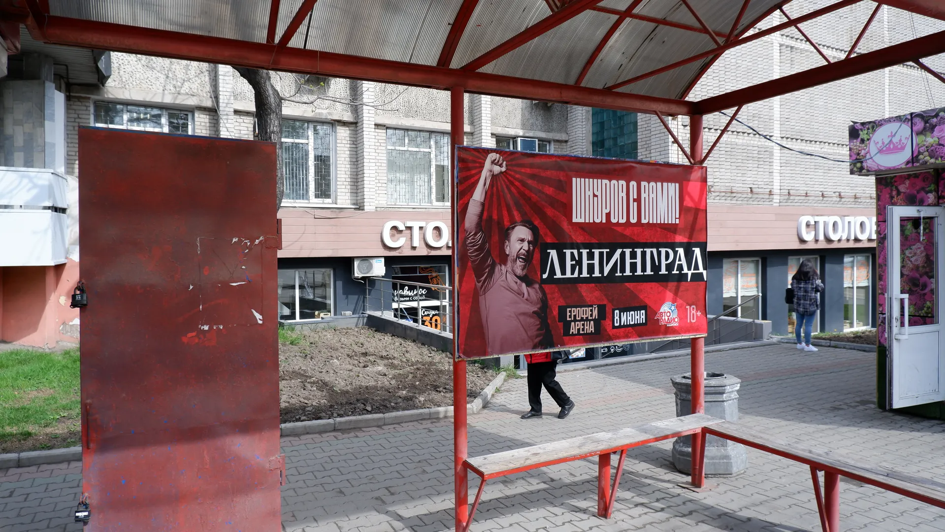 A red billboard shows a person raising a fist, with buildings and shops in the background. Next to the billboard is a red door and a bench. A person is walking past the billboard.