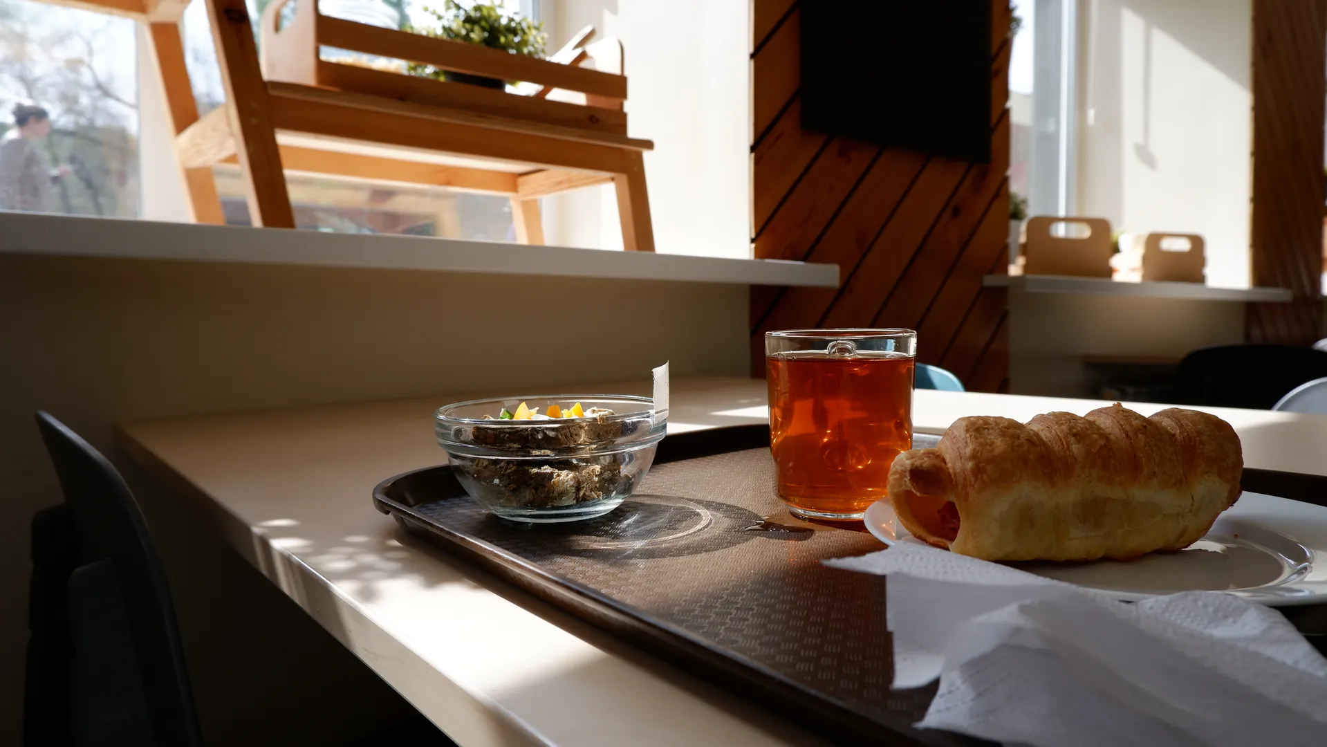 A tray on the table holds a teacup, a croissant, and a bowl of food. Sunlight shines through the window onto the table, with wooden chairs and a wall in the background.