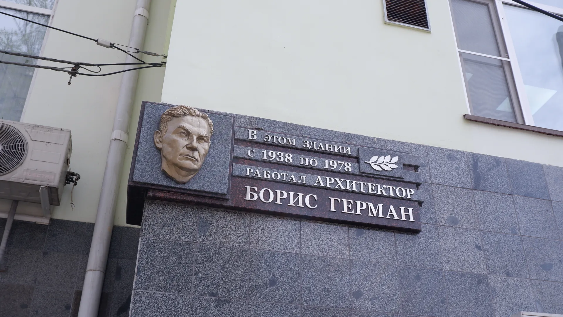A commemorative plaque on a wall, featuring a person’s relief and Russian text. To the left of the plaque is an air conditioning unit, and to the right is a window. The wall is made of gray tiles.