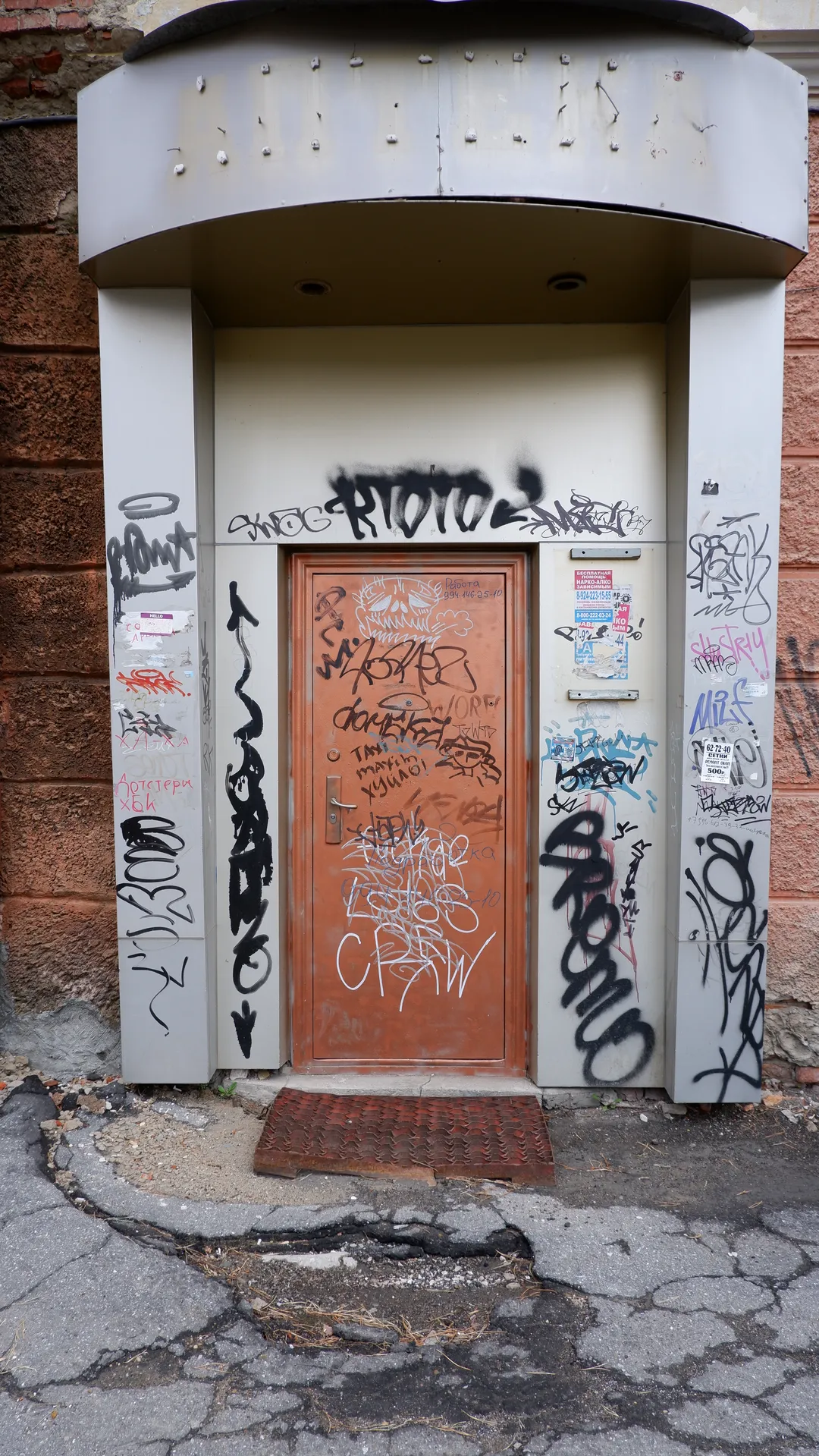 A door and surrounding walls covered in graffiti. Above the door is an arched structure with columns on both sides. The ground is cracked and damaged.