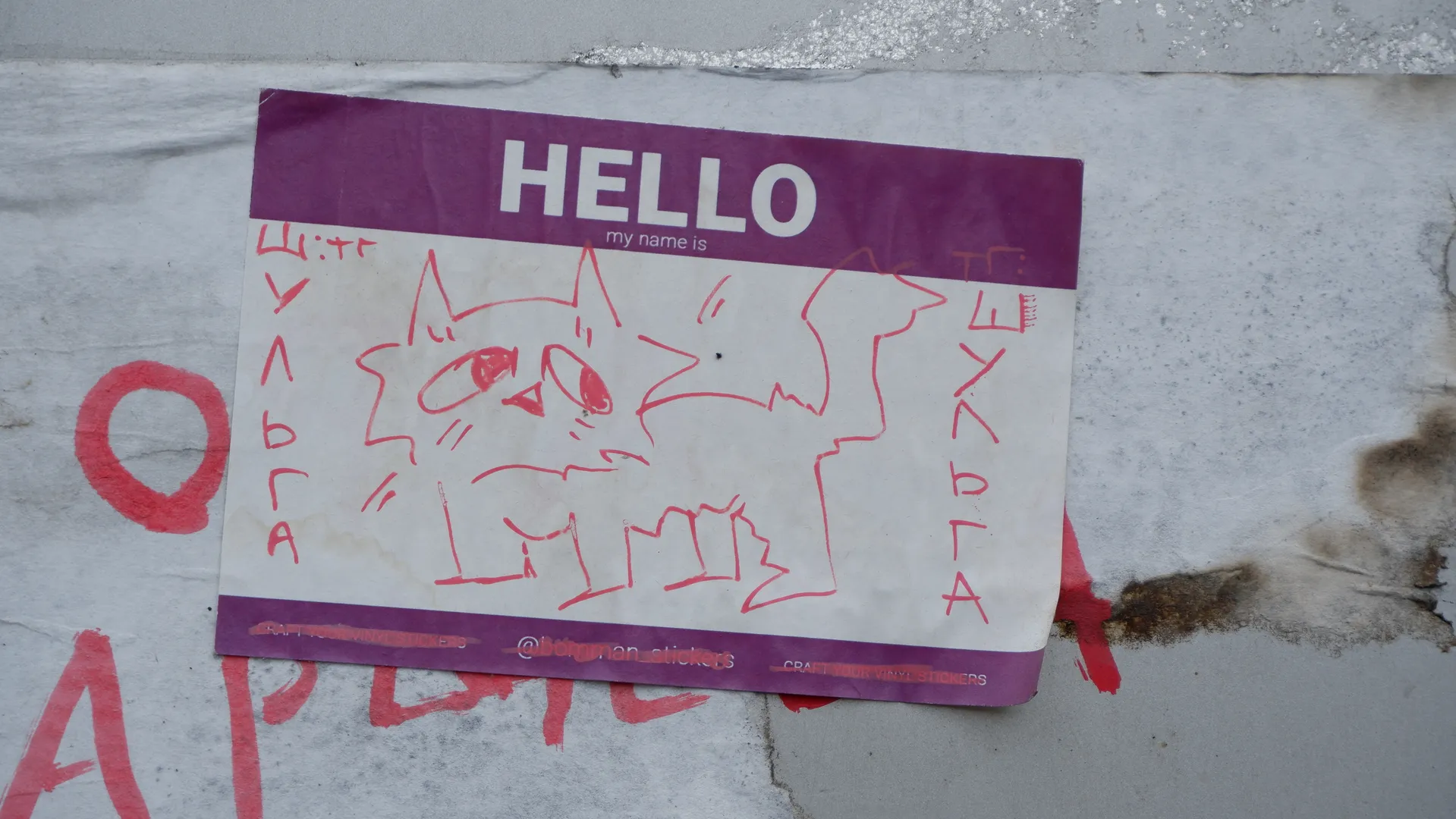 A piece of paper on the wall shows a red line-drawn cat with the text “HELLO my name is”. Red graffiti surrounds the paper, and the background is a gray wall.