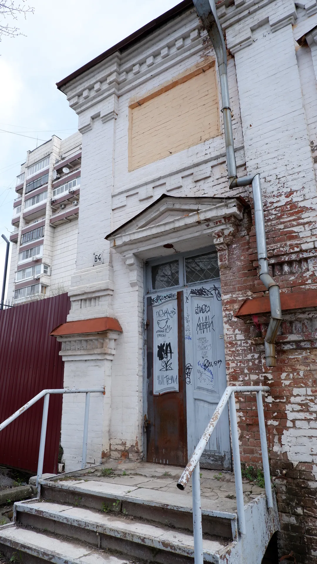 At the entrance of a white building are steps and handrails, with graffiti on the door. Some bricks on the exterior wall are exposed, and there are pipes on the right. A high-rise building is visible in the background.
