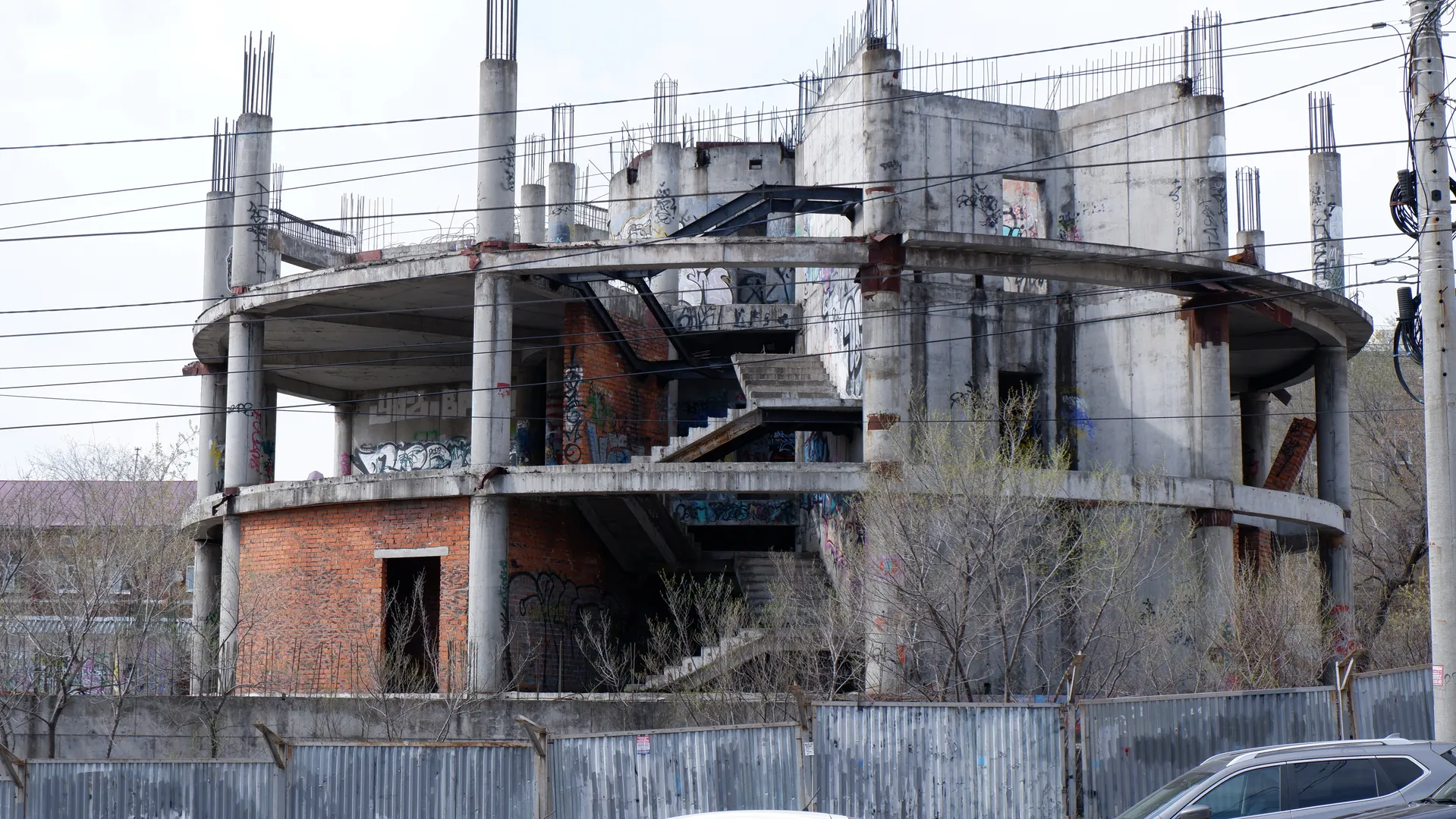 An unfinished building with exposed structure and graffiti on the walls. A fence stands in front, with trees surrounding the area. The sky is overcast.
