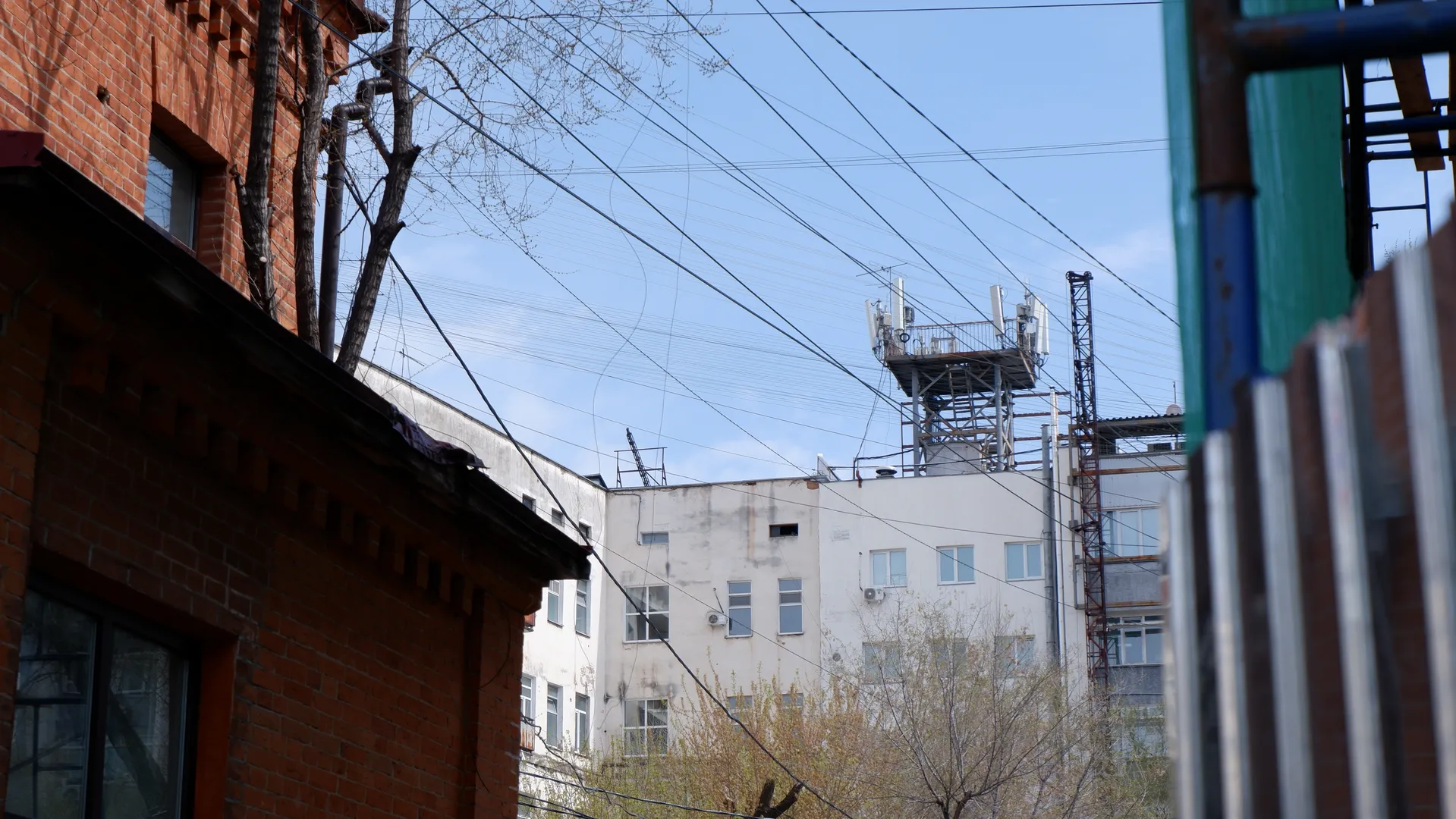 Next to a red brick building is a white one, with a communication tower on the roof. Many power lines cross the sky, and metal railings are in the foreground.