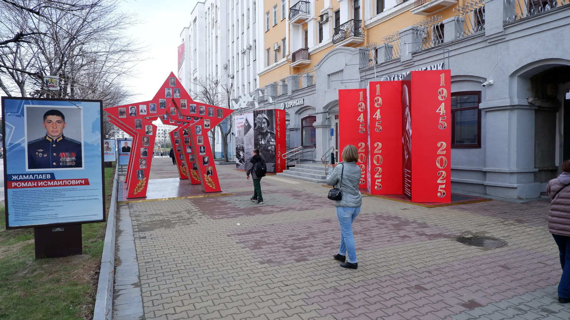 Several people are on the street with red decorations and posters nearby. The posters show soldiers’ photos and information, and the decorations display the years “1945” and “2025”. In the background are multi-story buildings.