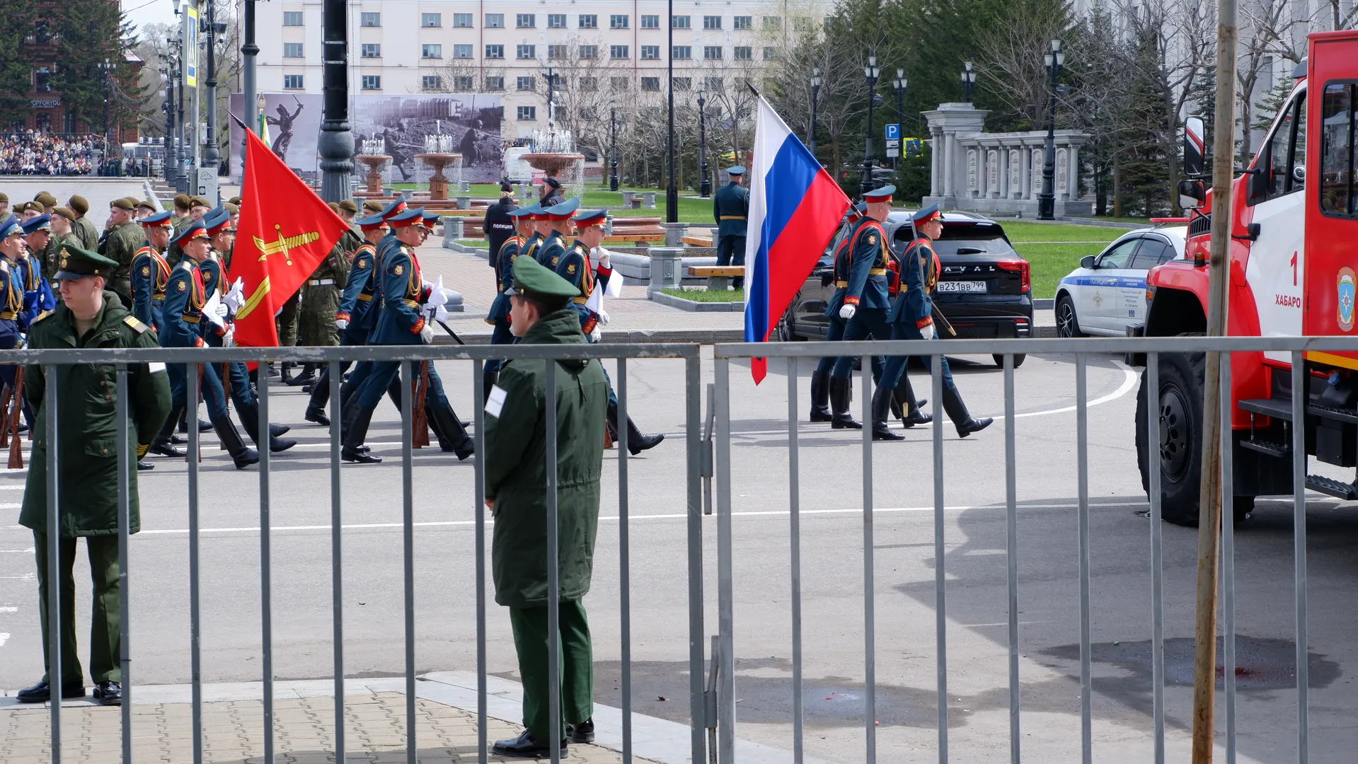 Soldiers marching in neat formation, holding red and tricolor flags of blue, white, and red. In the foreground, two personnel in green uniforms stand, with several cars parked in the background.