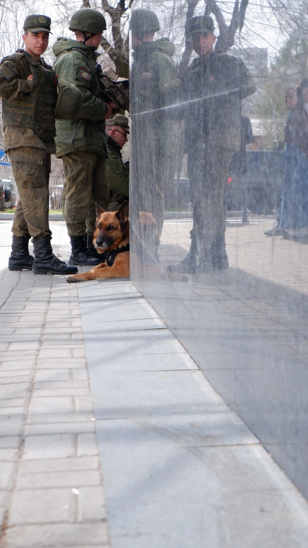 Several soldiers stand beside a statue, with a German Shepherd lying on the ground. Trees and pedestrians appear in the background.