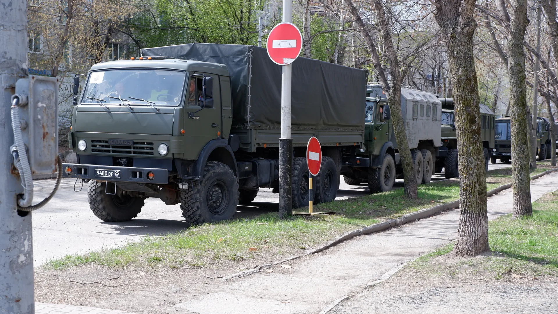 Several military trucks parked by the roadside, painted green and covered with black tarps. Traffic signs and trees are nearby. The road is concrete.