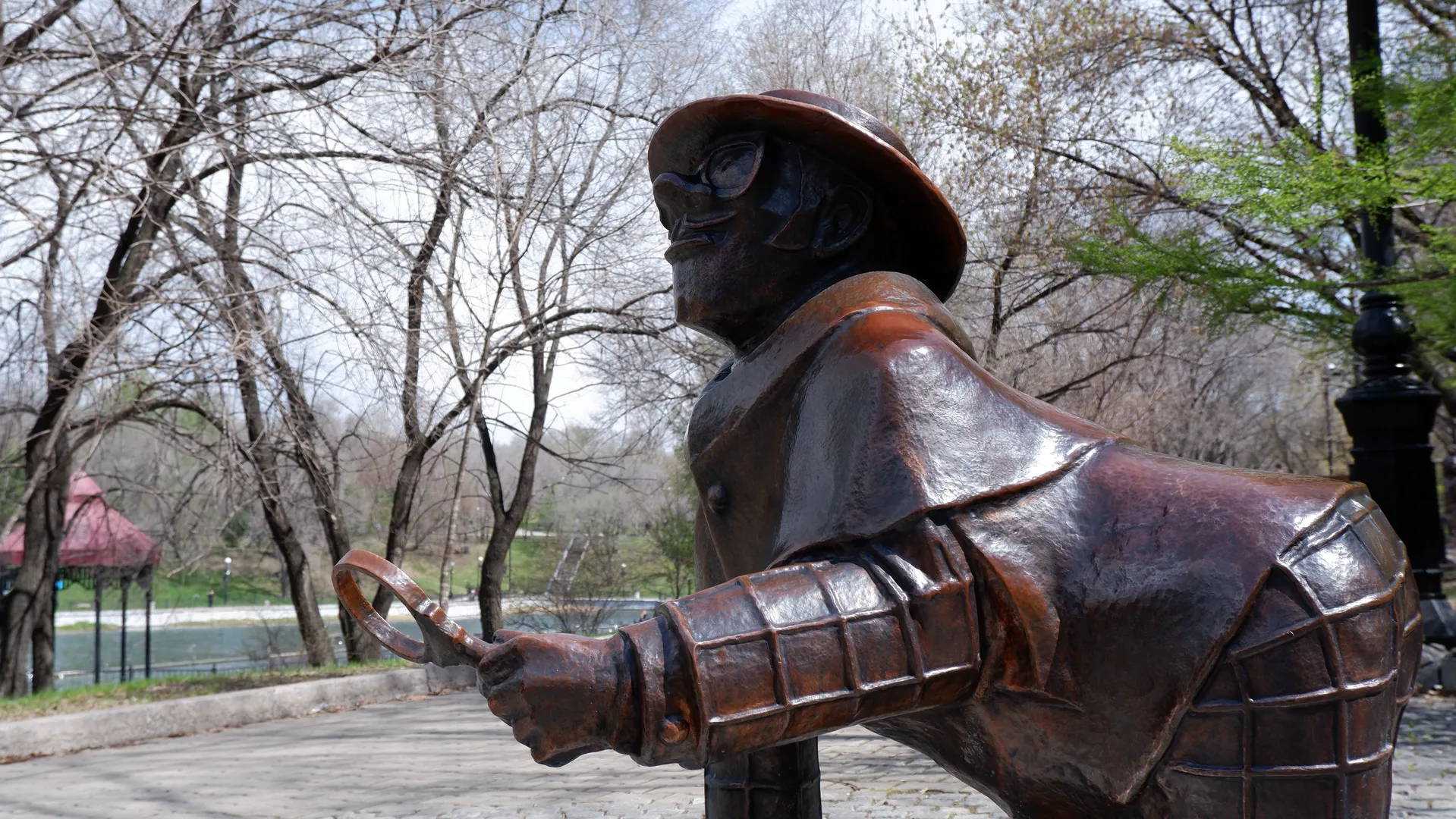 A sculpture depicts a person wearing a hat and holding a tool. Trees and distant buildings are in the background. The ground is paved with stone slabs.