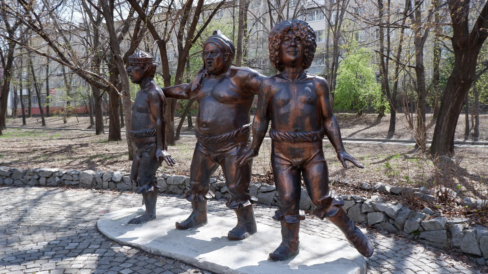 Three sculptures stand together in different poses. Trees and buildings are in the background. The ground is paved with stone slabs and surrounded by stone walls.