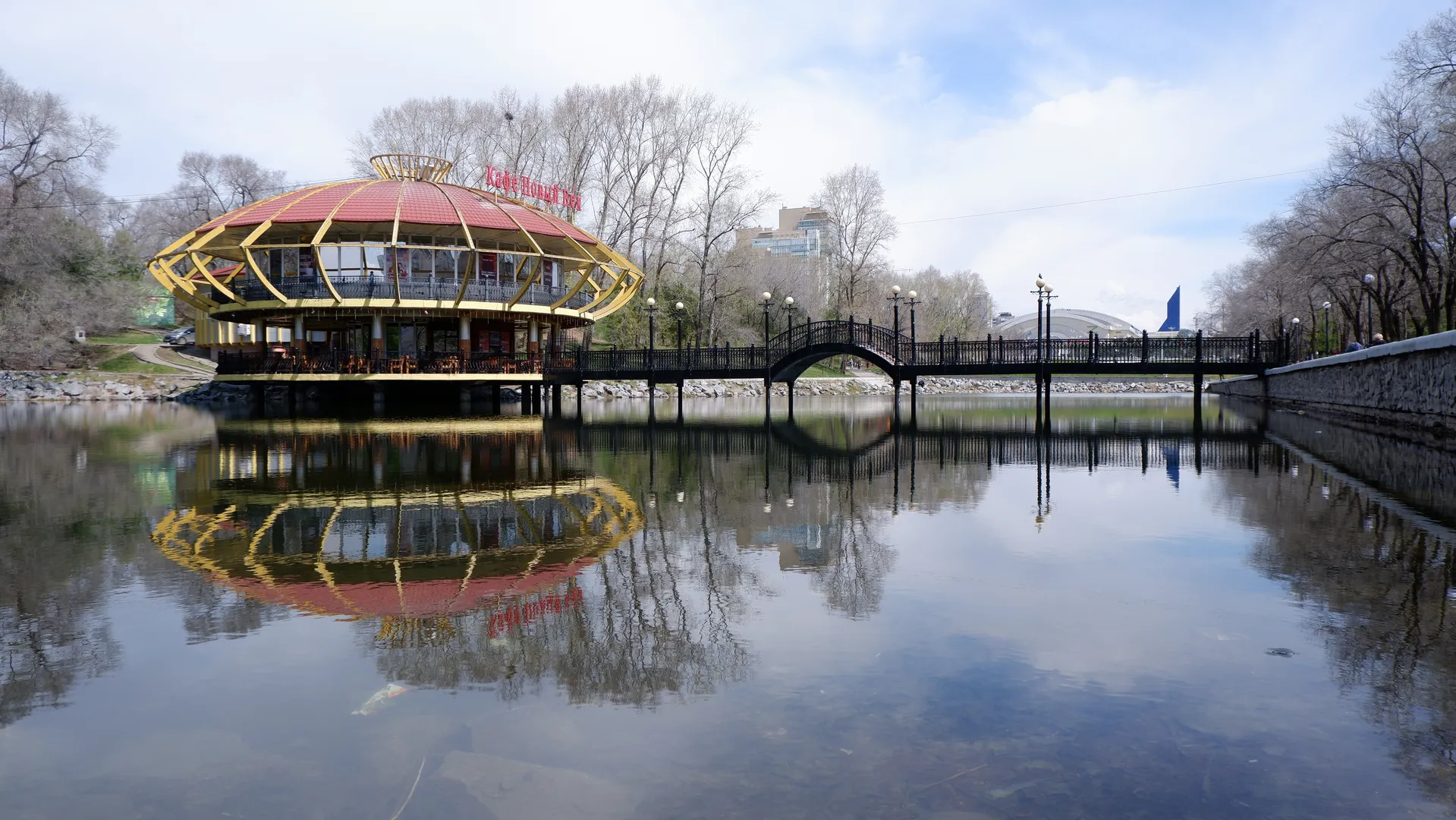 A two-story building sits on the water, with a red roof and yellow framework. In front of the building is an arched bridge connecting both sides of the bank. The calm water reflects the building and the bridge. The surroundings include bare trees and distant buildings.