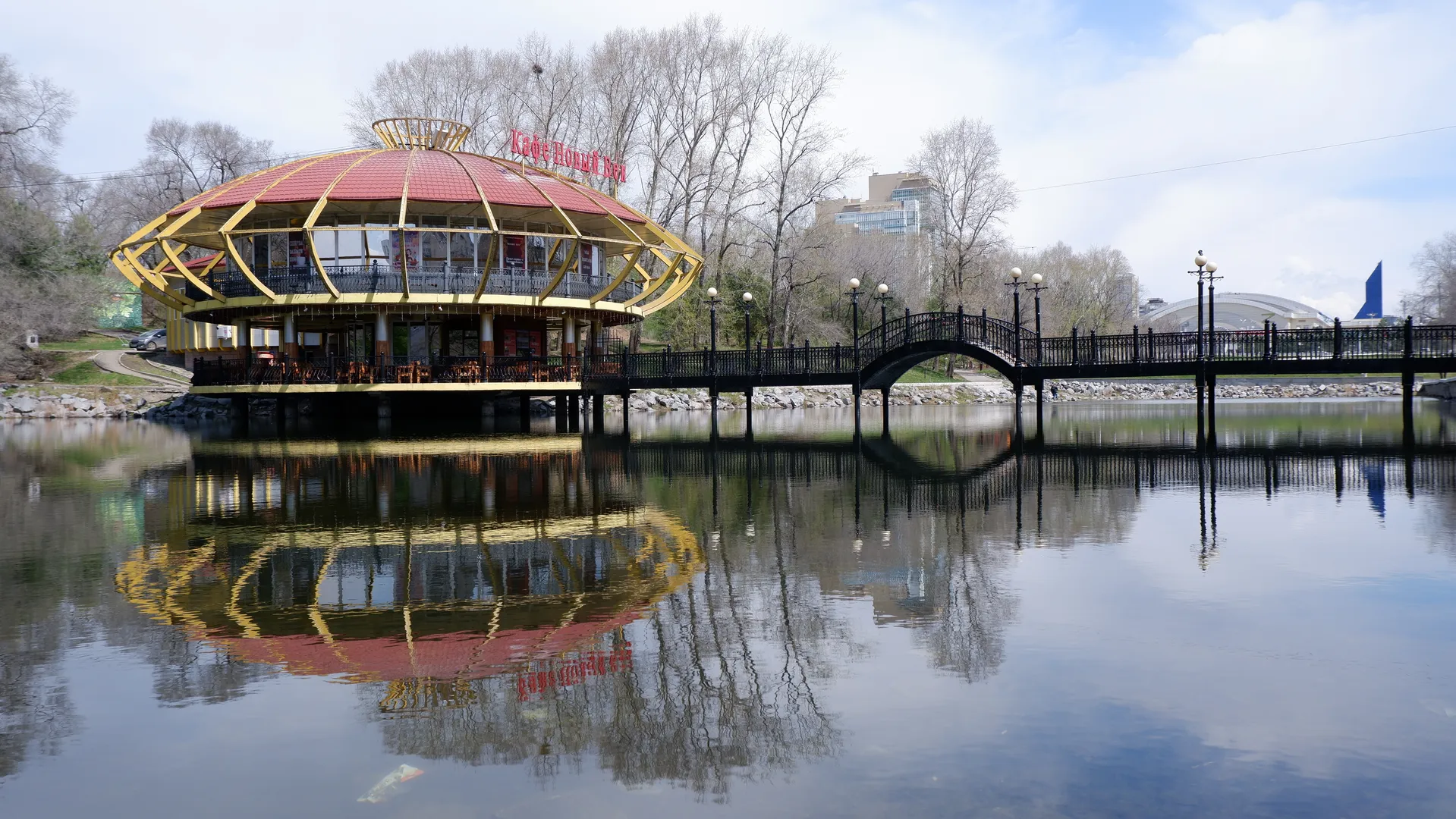 A two-story building sits on the water, with a red roof and yellow framework. In front of the building is an arched bridge connecting both sides of the bank. The calm water reflects the building and the bridge. The surroundings include bare trees and distant buildings.