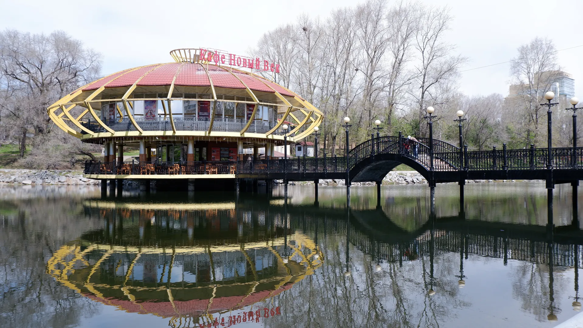 A two-story building sits on the water, with a red roof and yellow framework. In front of the building is an arched bridge connecting both sides of the bank. The calm water reflects the building and the bridge. The surroundings include bare trees and distant buildings.