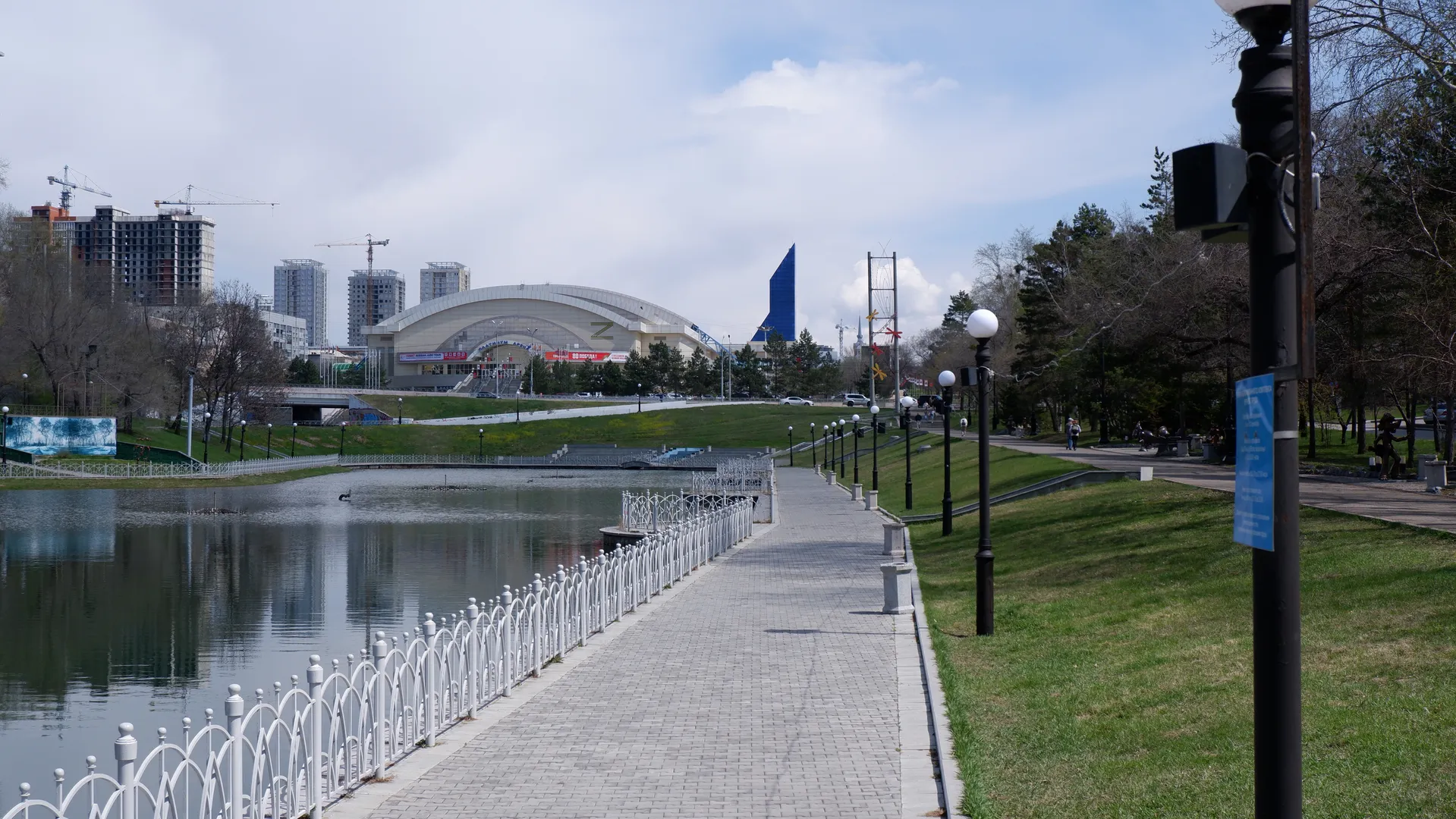 A stone-paved path runs along the water, with white railings and street lamps on both sides. In the distance stands a large building with a curved roof, and in front of it are green lawns and trees. There are few clouds in the sky and the weather is sunny.