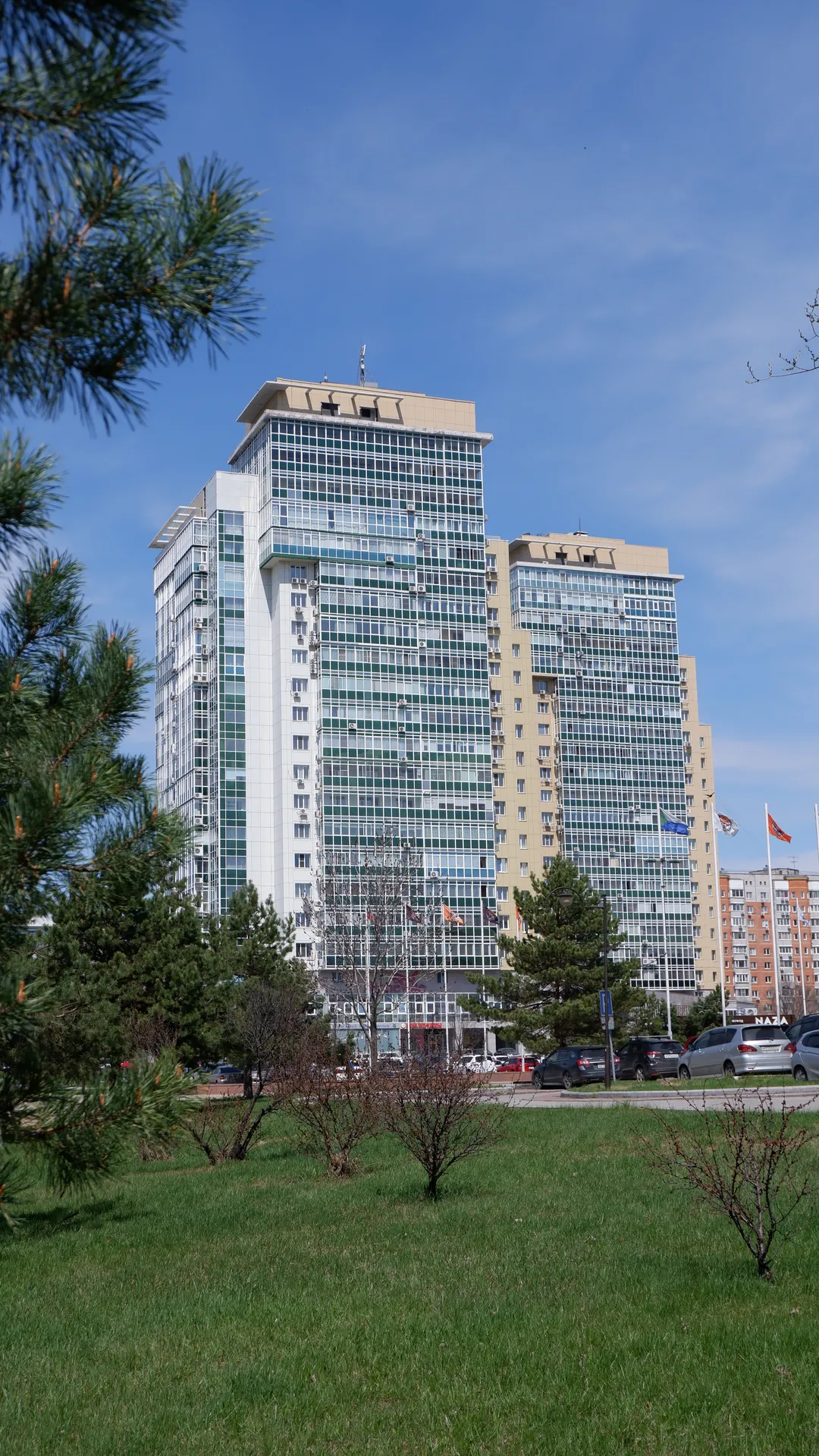 Several high-rise buildings stand tall under the blue sky, with facades mainly made of glass and concrete. In the foreground are trees and grass, and some vehicles are parked at the base of the buildings. The sky is clear with no clouds.
