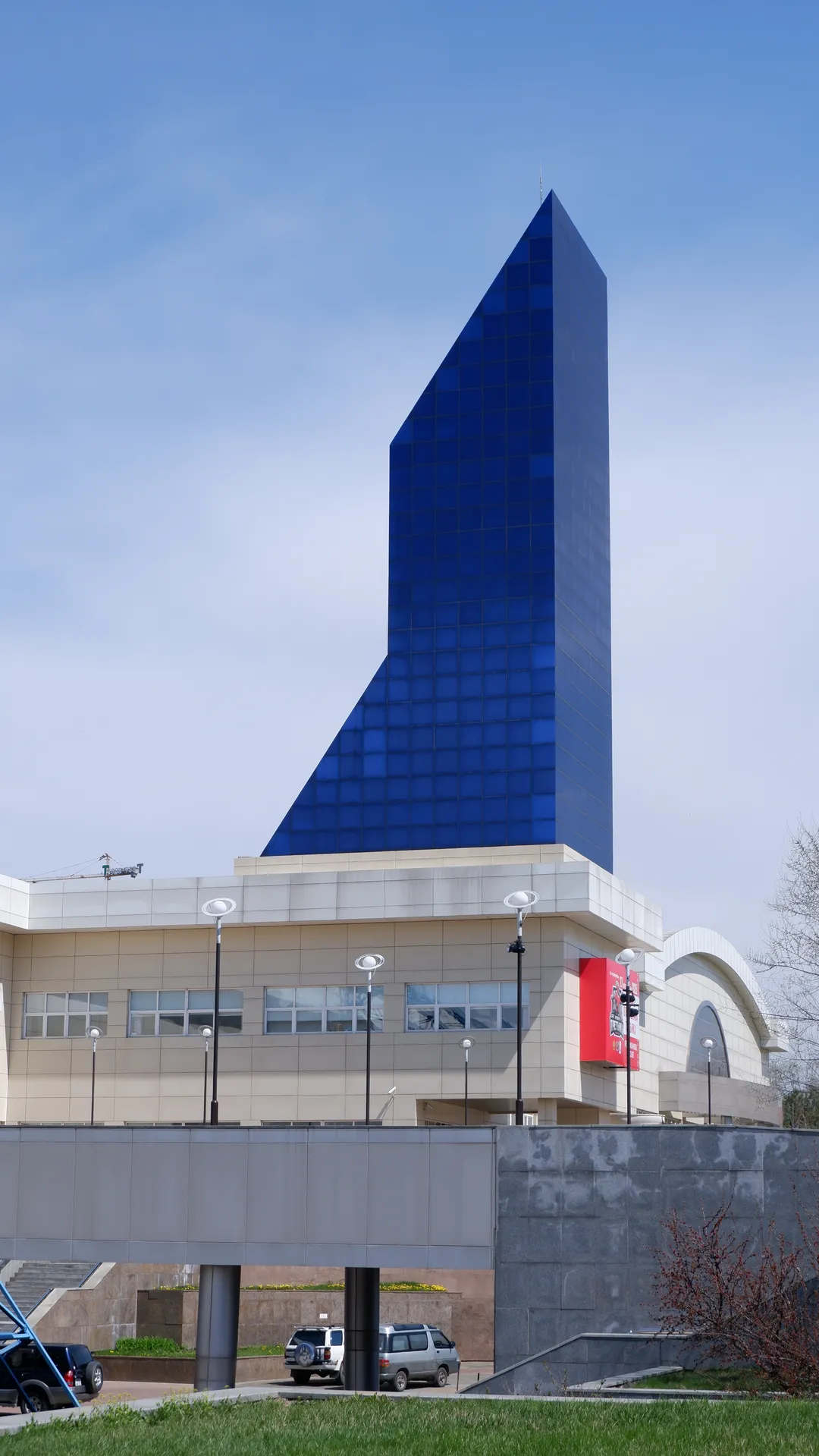 A modern building features a blue glass pyramid-shaped tower. The base of the building is made of light-colored stone, with several street lamps and a billboard in front. The sky is clear, with some trees and shrubs in the background.