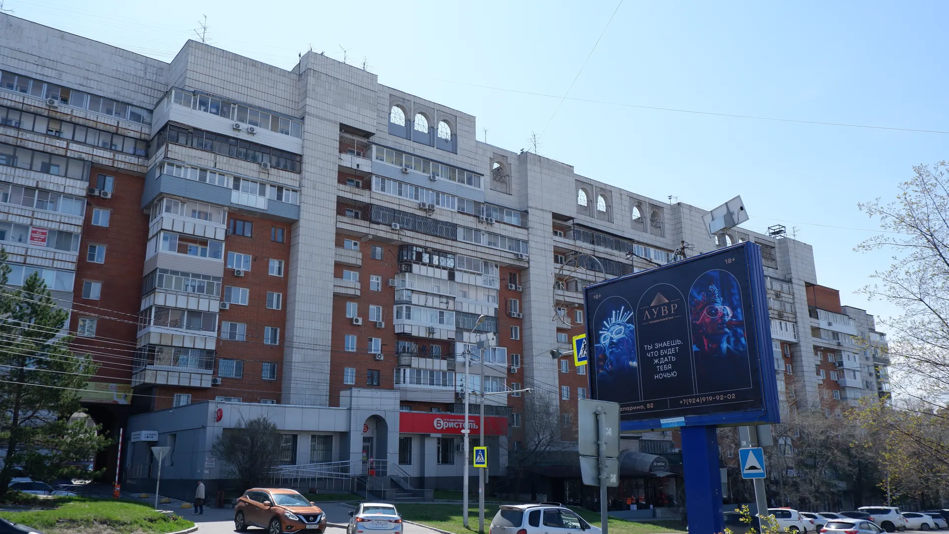 A multi-story residential building with a brick and concrete exterior and neatly arranged windows. In front of the building is a large billboard with text and images. Several cars are driving on the street, with pedestrians and trees nearby.