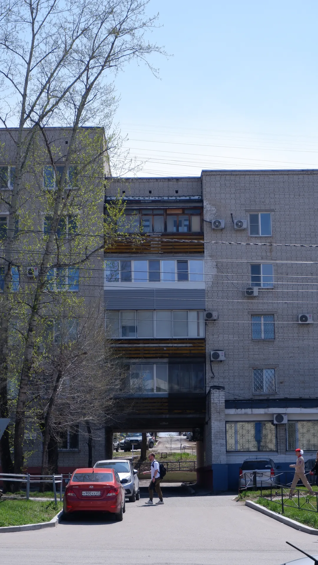A multi-story residential building with a brick and concrete exterior and neatly arranged windows. In front of the building is a parking lot with several parked cars. A few people are walking in front of the building, with power lines and trees in the background.